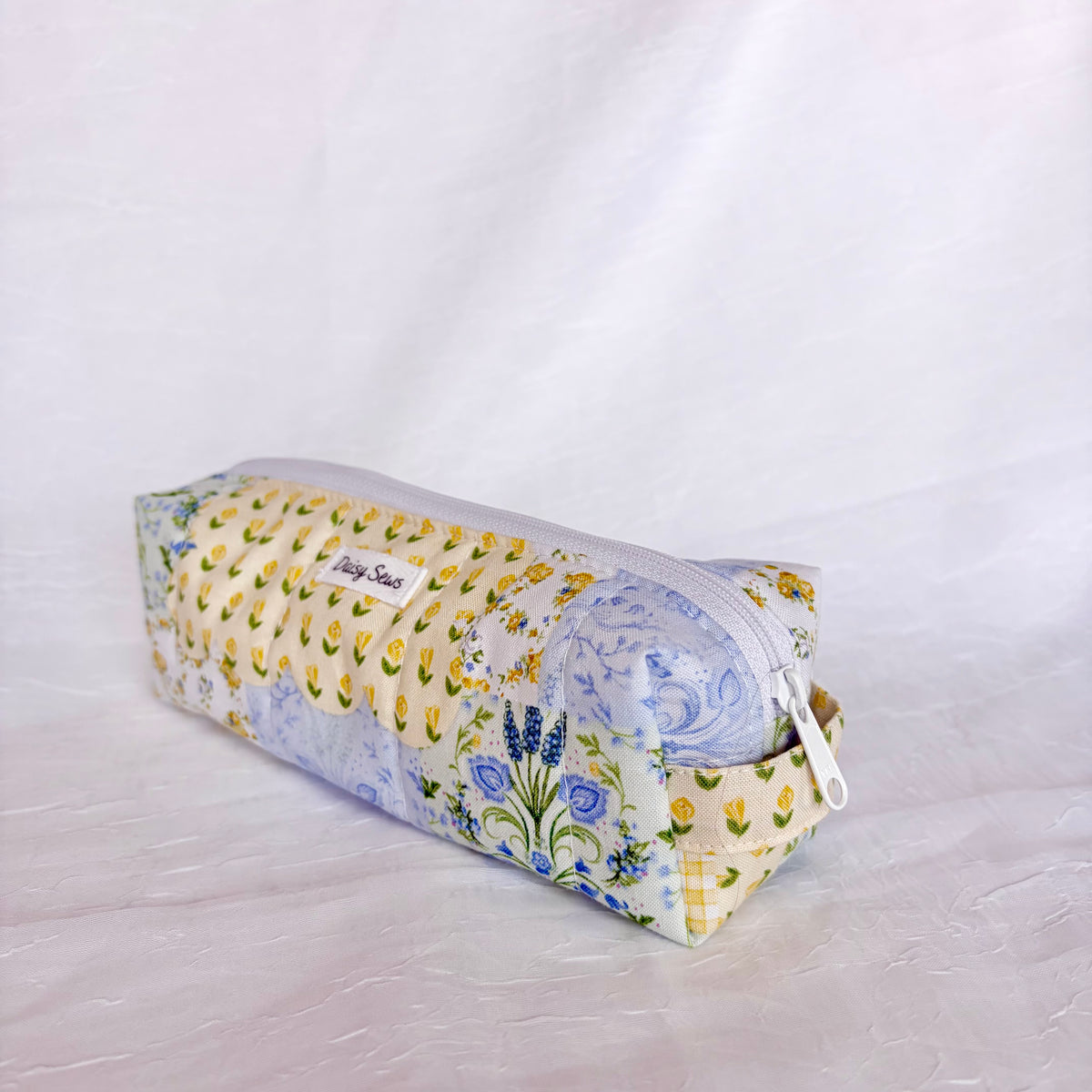 A rectangular zippered pouch with a multicolor floral and botanical print, featuring yellow tulips, blue flowers, and green leaves on a white background, with a white zipper pull and fabric handle.
