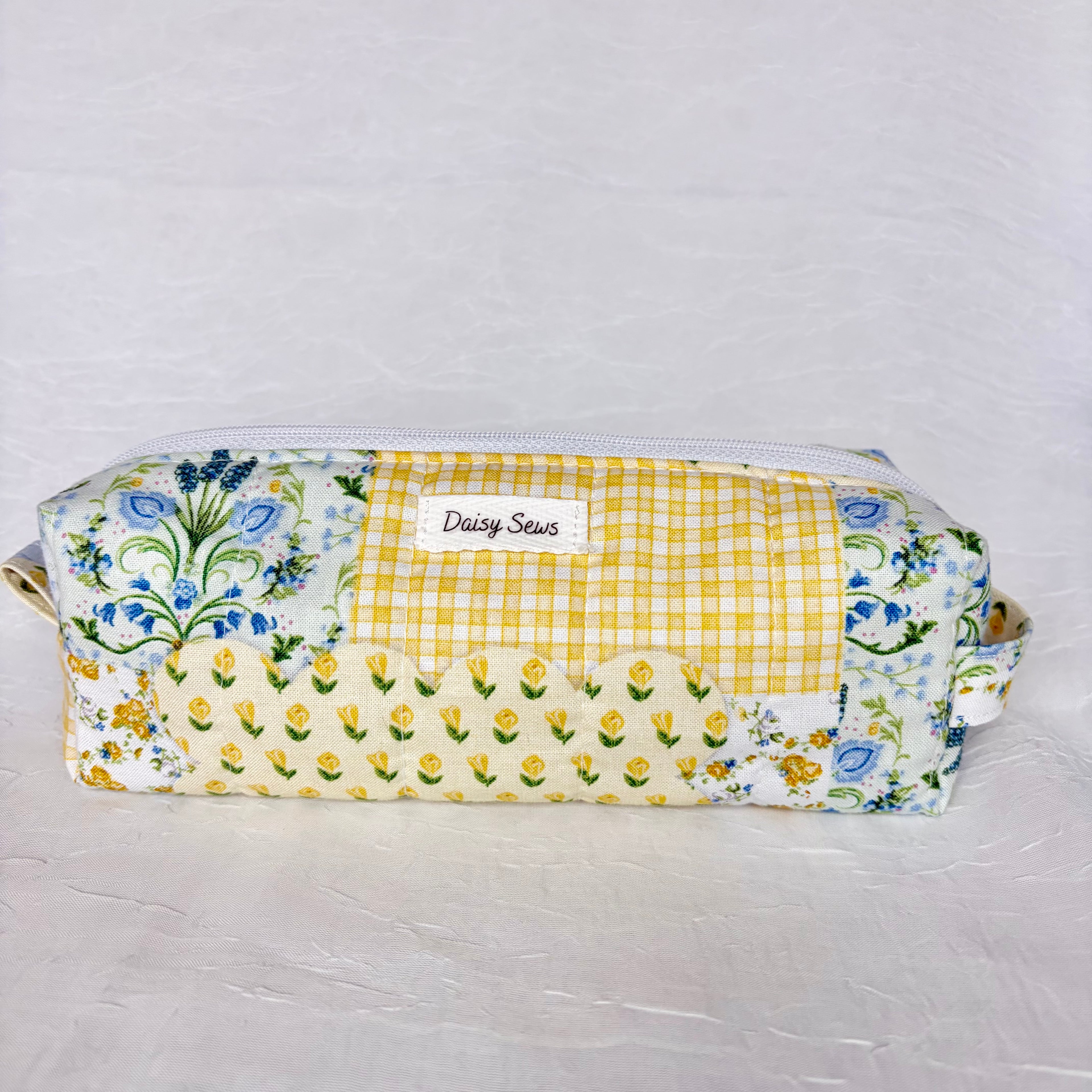 Blue and yellow floral patchwork boxy pencil bag with white zipper and a white label on white background