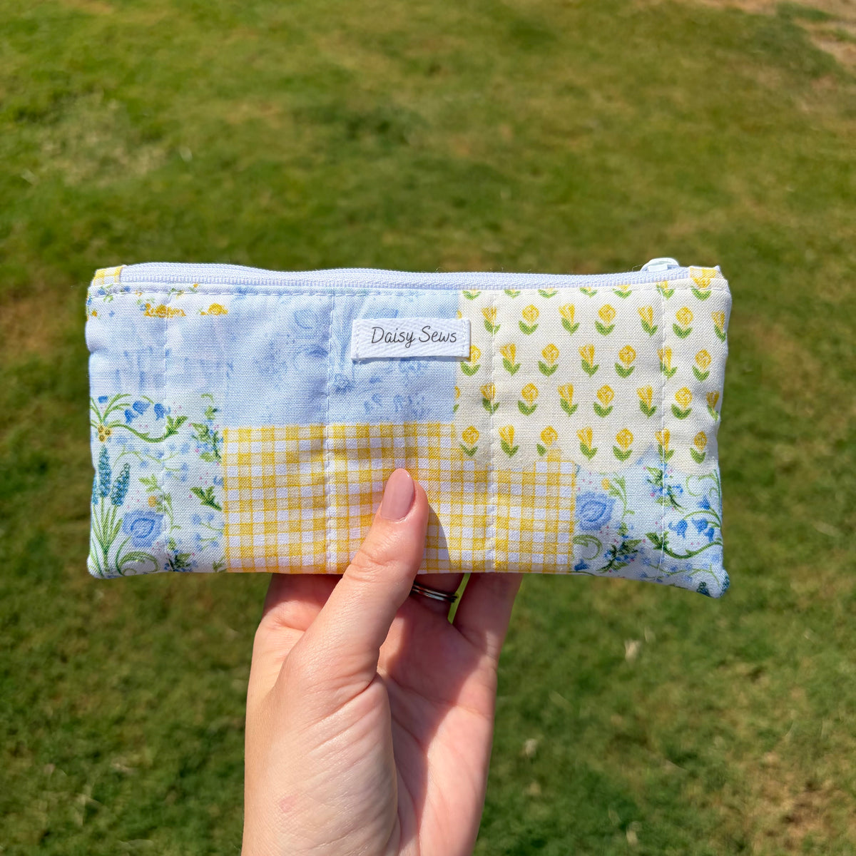A hand holds a small rectangular patchwork pouch outdoors on a grassy lawn. The pouch has a light blue floral print, a yellow gingham panel, a white floral print with yellow tulips, and a white label reading Daisy Sews.