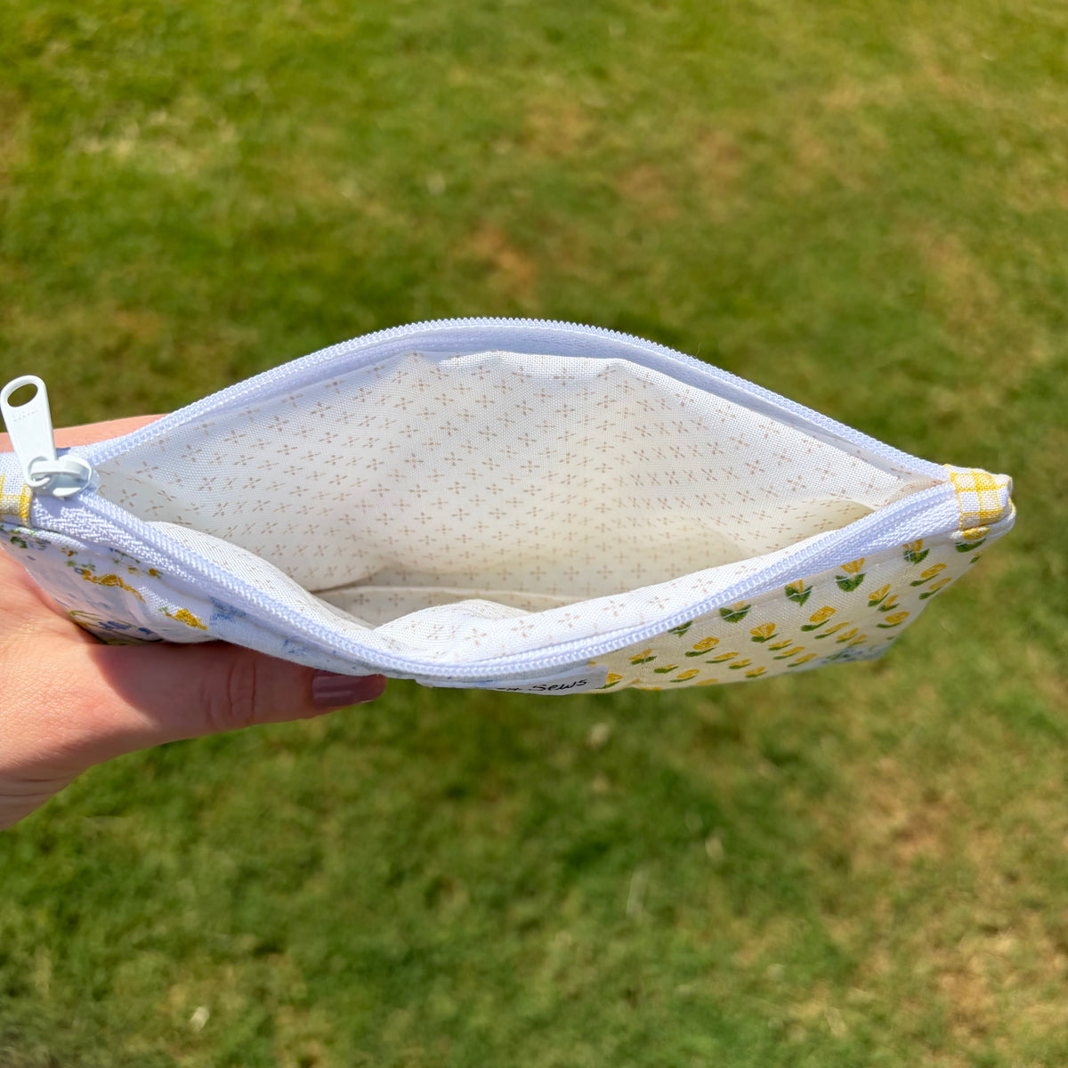 A hand holding open a small zip pouch outdoors over a grassy lawn. The pouch has a white interior with tiny blue dots and a white exterior printed with small yellow and green floral motifs.