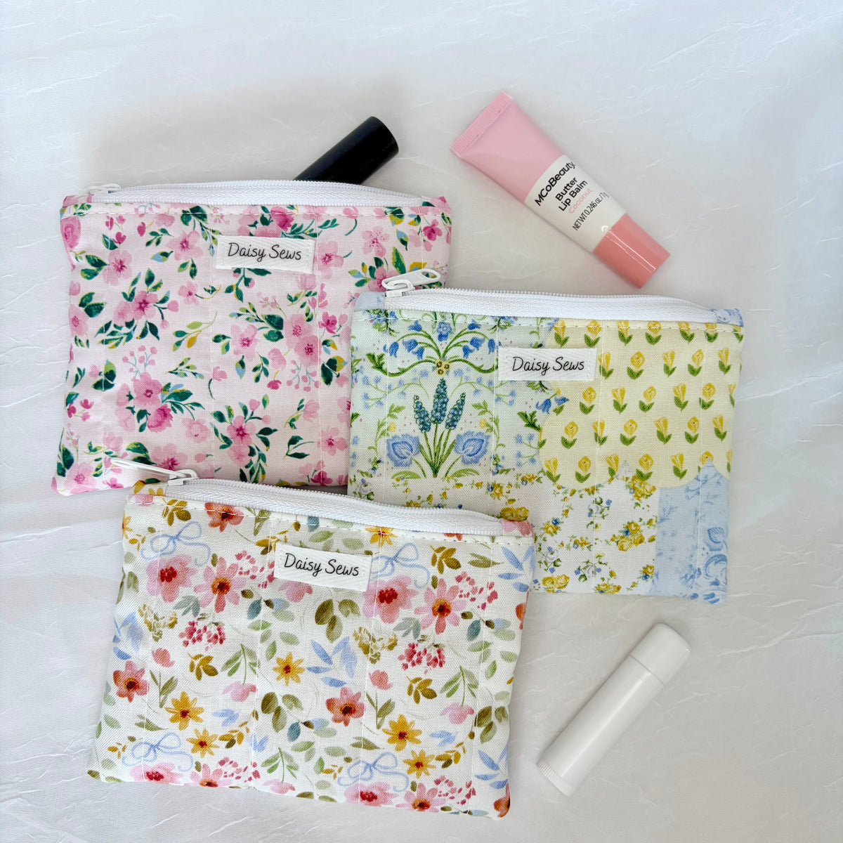 Three floral zippered cosmetic pouches by Daisy Sews are arranged on a light fabric background with small beauty props: a pink lip balm tube and a white lip balm stick. The pouches have different floral prints in pink, green, blue, and multicolor tones, and each has a visible Daisy Sewslabel.