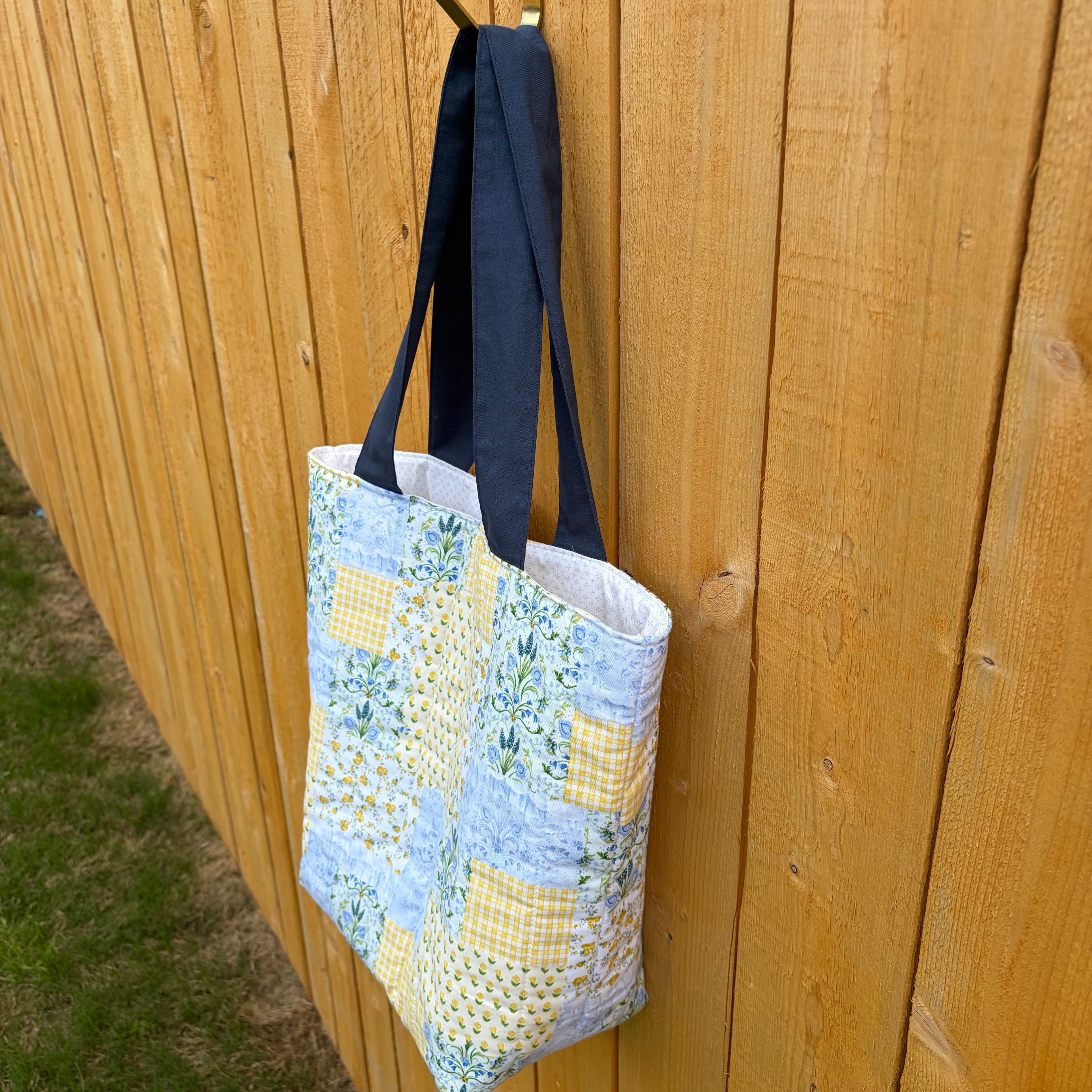 Side View of Blue and Yellow floral Patchwork Tote Bag with navy handles hanging on wooden fence