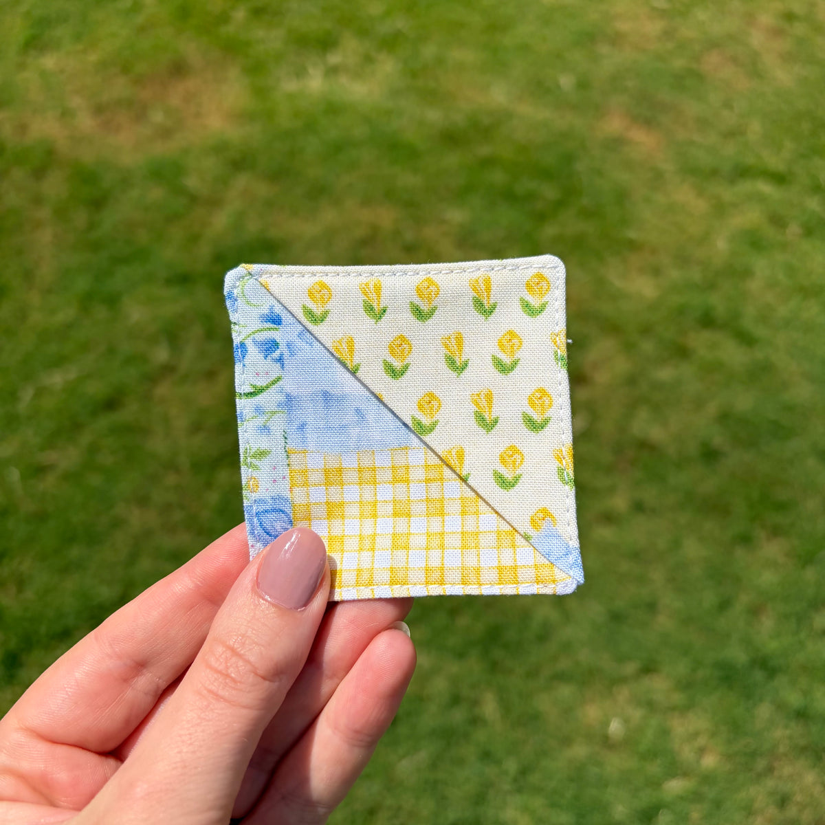 A hand with mauve-painted nails holds a small square fabric patch with three patterned sections: yellow tulips, blue flowers on white, and a yellow gingham check. The background is a blurred green grass lawn.