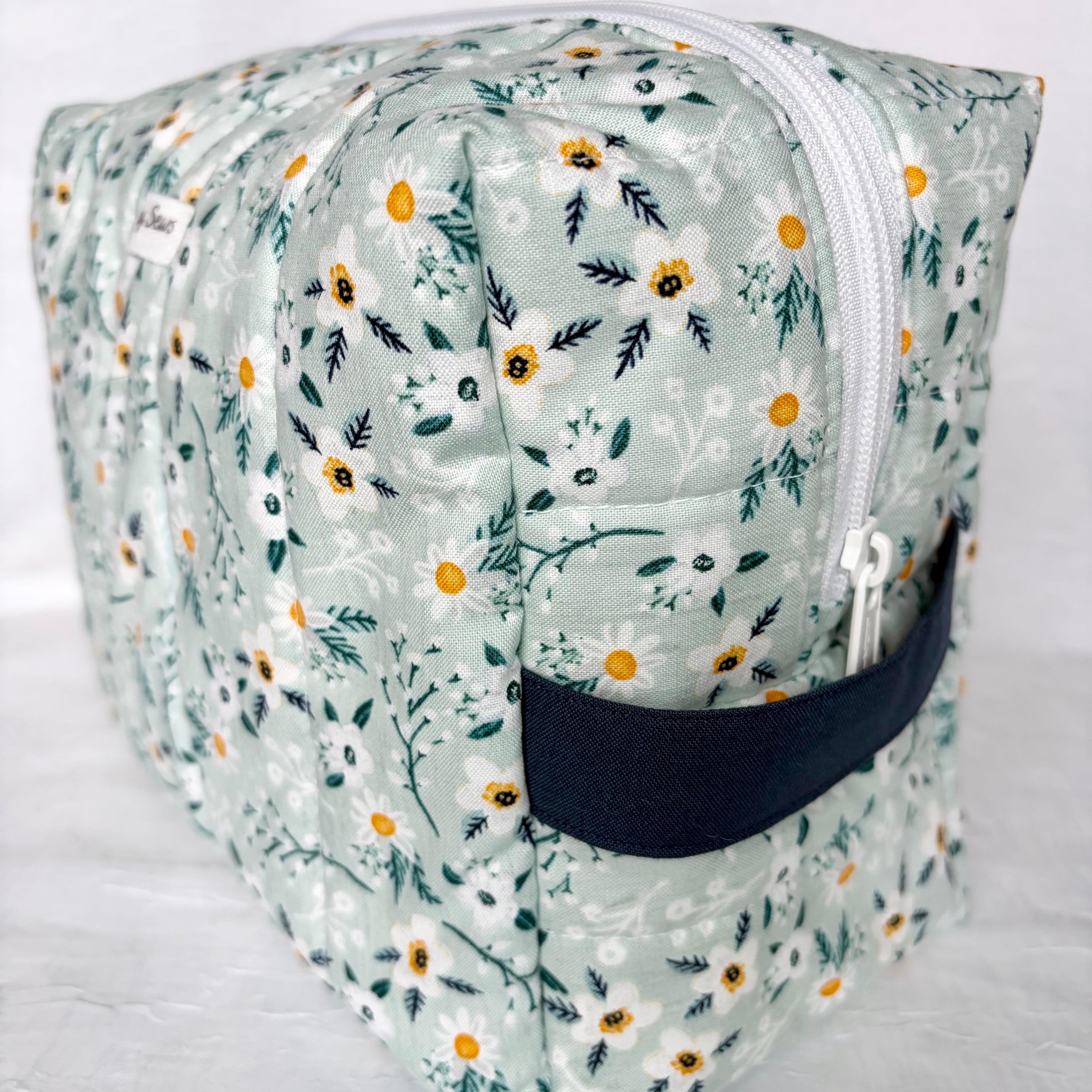 side view of navy handle and white zipper on blue with white flowers and teal leaf patterned boxy bag with white label on white background