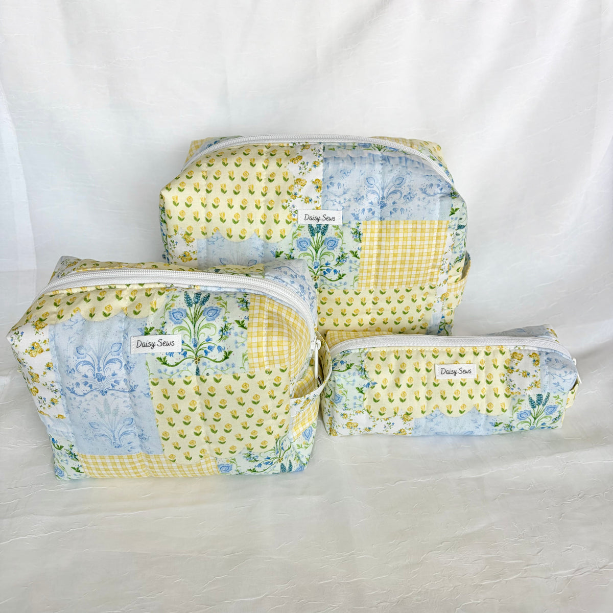 Three patchwork zippered cosmetic bags in yellow, blue, and white fabrics, labeled 'Daisy Sews' on a white fabric backdrop.