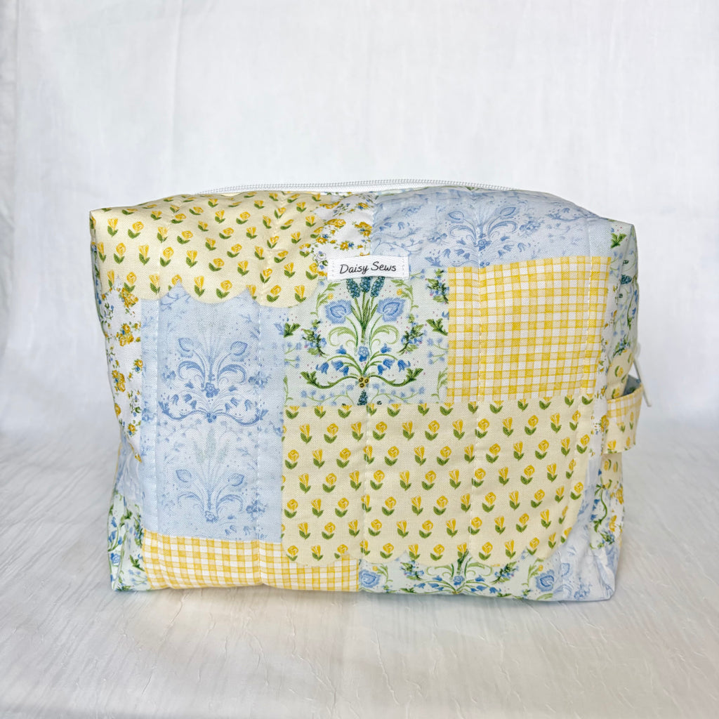 A patchwork fabric cosmetic pouch with a yellow and blue floral pattern, featuring a small white label that reads 'Daisy Sews'.