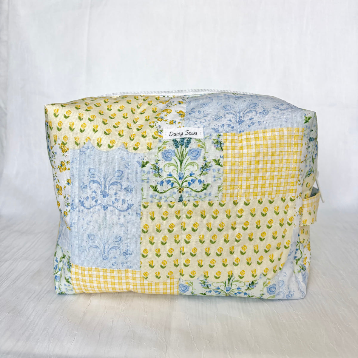A patchwork fabric cosmetic pouch with a yellow and blue floral pattern, featuring a small white label that reads 'Daisy Sews'.