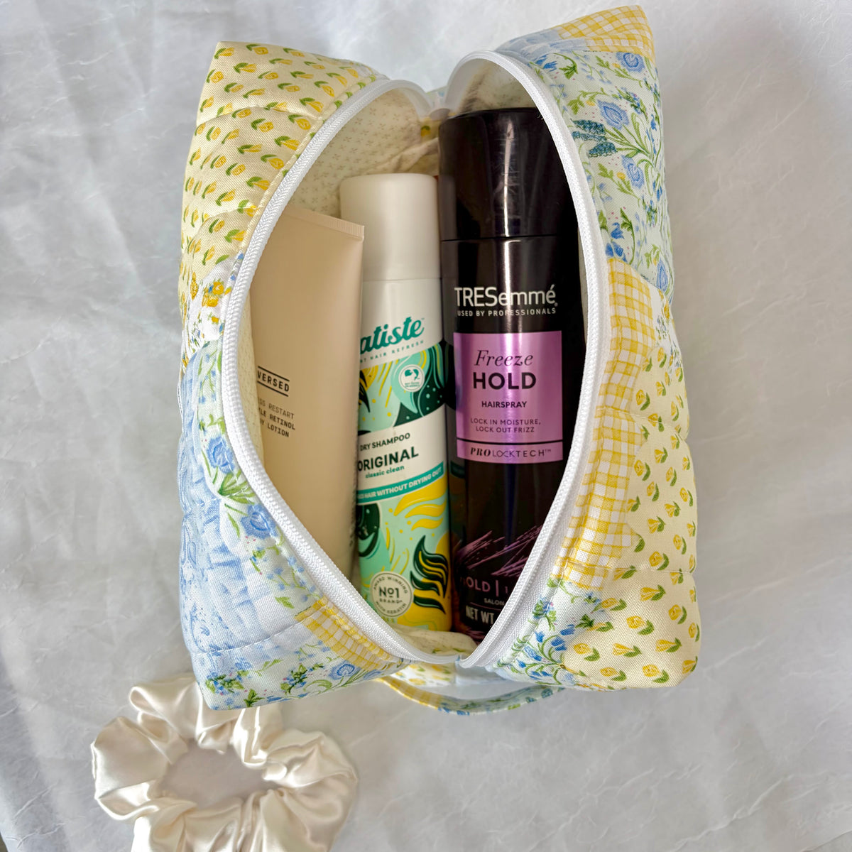 A fabric zippered pouch with a yellow and blue floral print sits open on a white textured fabric background, containing personal care items including TRESemmé hair spray and  lotion. A matching scrunchie is placed below the pouch.