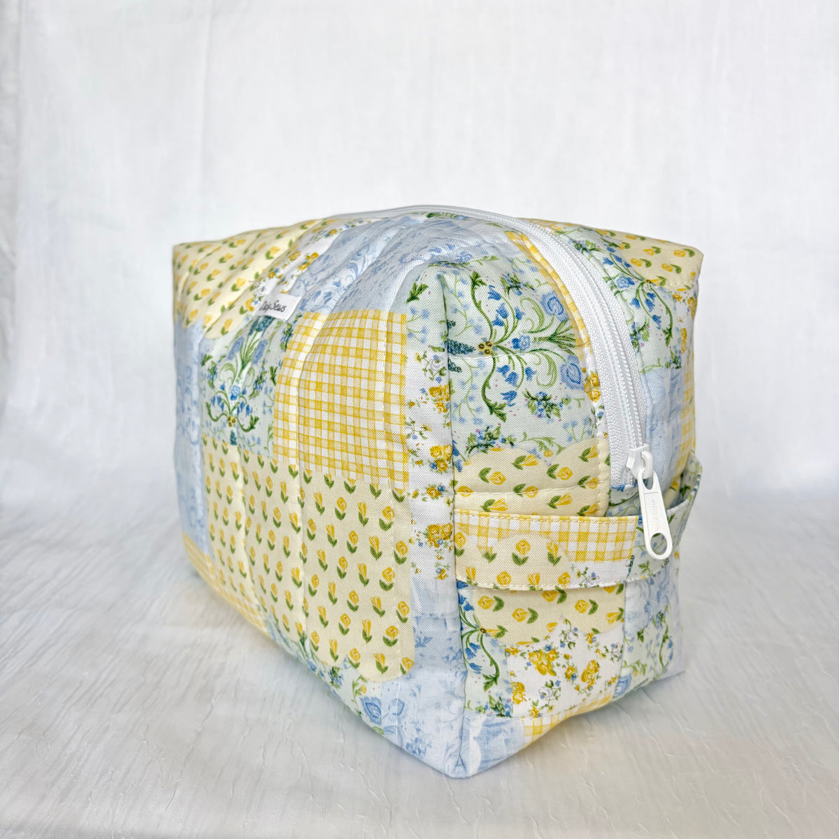 A square patchwork zippered pouch made from various light-colored fabrics, including yellow floral prints, blue floral prints, and yellow gingham, set against a white fabric backdrop.