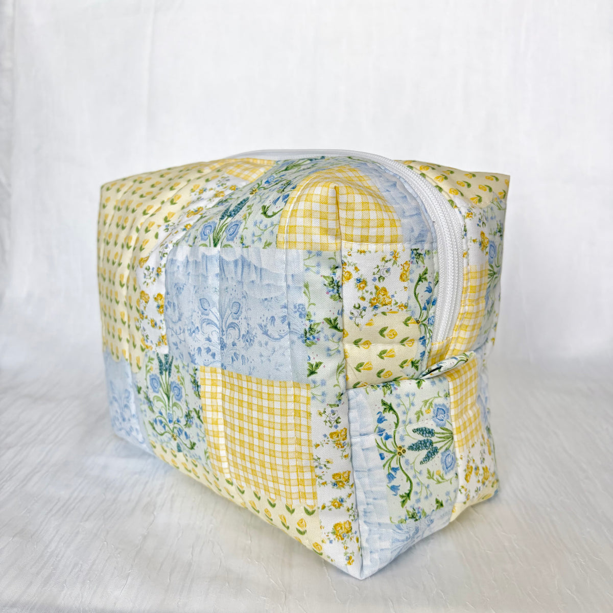 A square patchwork quilted cosmetic bag with a zipper closure, featuring yellow gingham, blue floral, and light blue floral patterns, set against a white fabric backdrop.