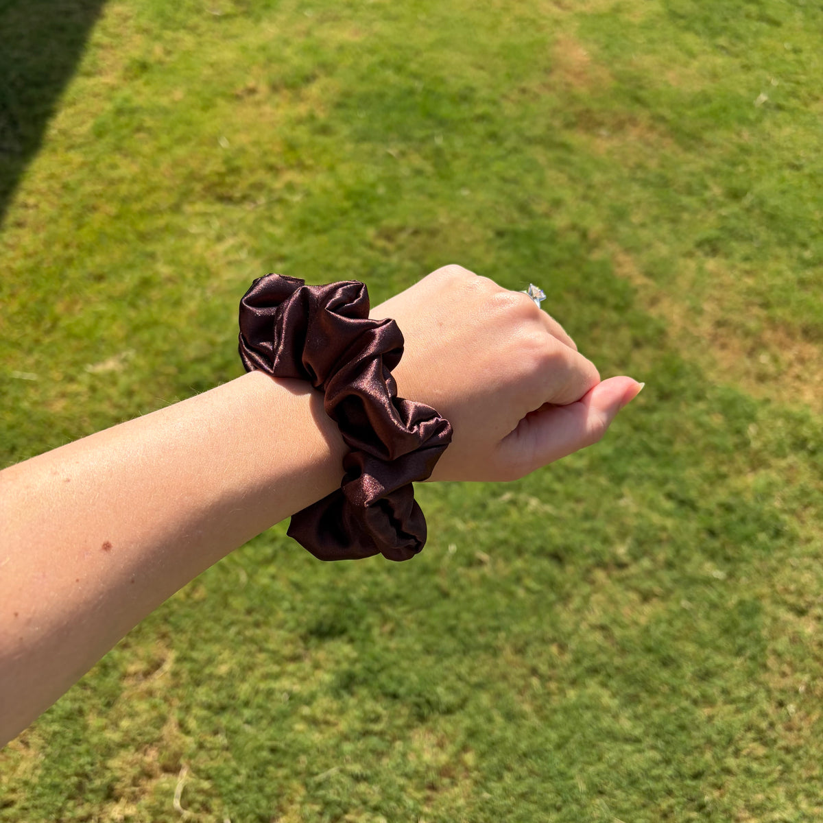A dark brown satin scrunchie worn on a person's wrist, photographed outdoors against a blurred green grass background.