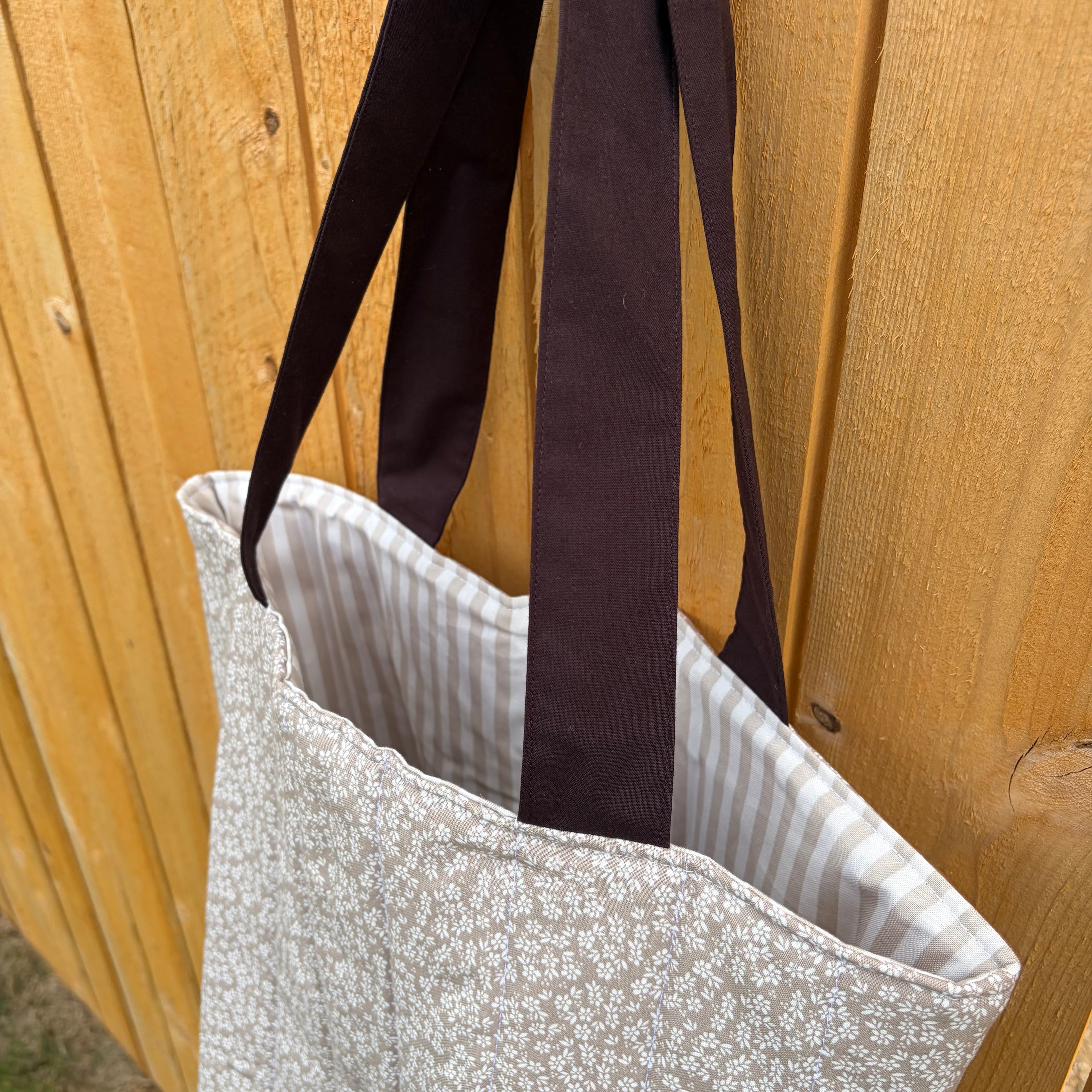 close up side view of Brown floral tote bag with dark brown handles and tan vertical striped interior hanging on a wooden fence