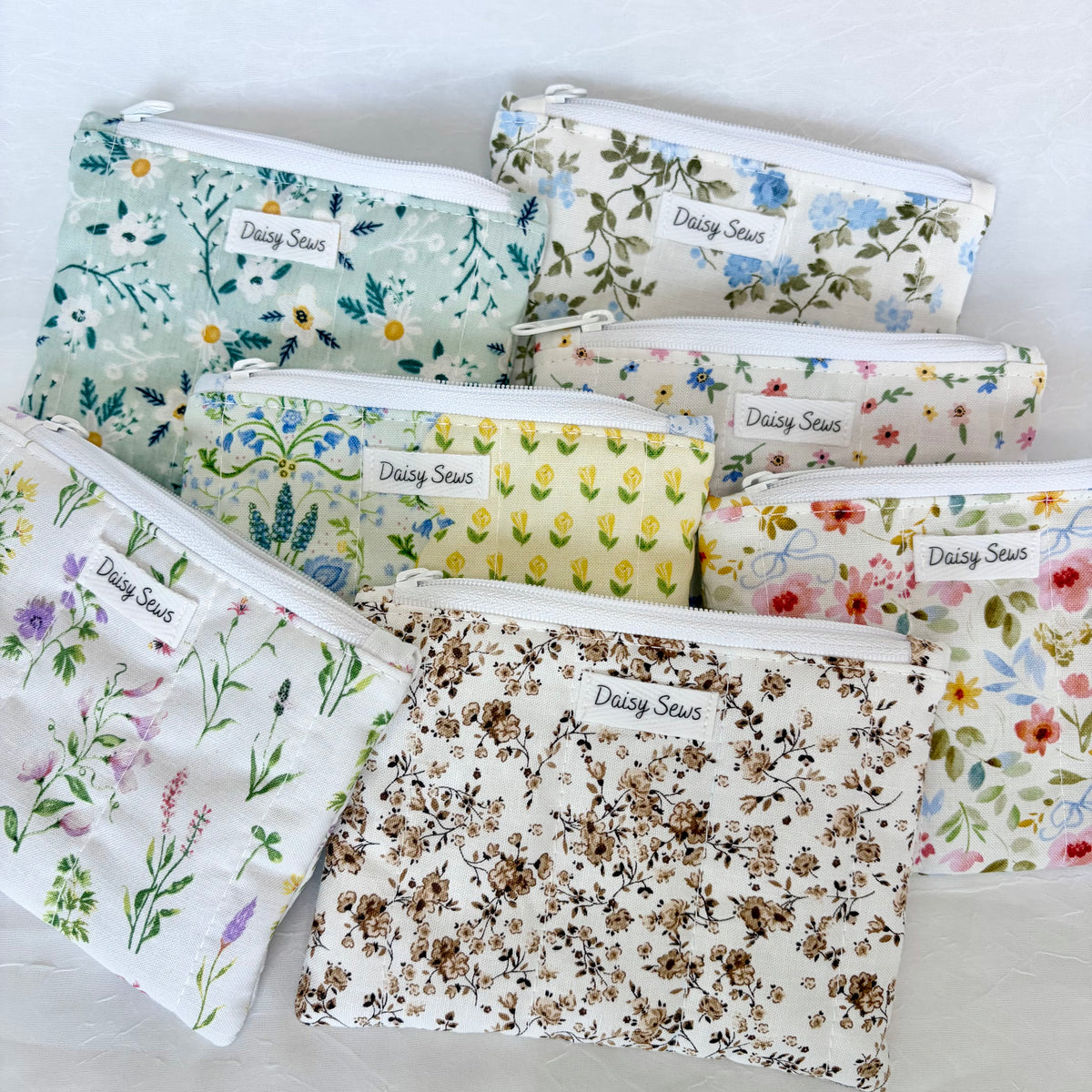 Seven fabric zip pouches with white zippers and 'Daisy Sews' labels, arranged in a flat-lay on a white textured background. Each pouch features a different floral and botanical print in soft pastel colors including green, yellow, pink, blue, and brown.
