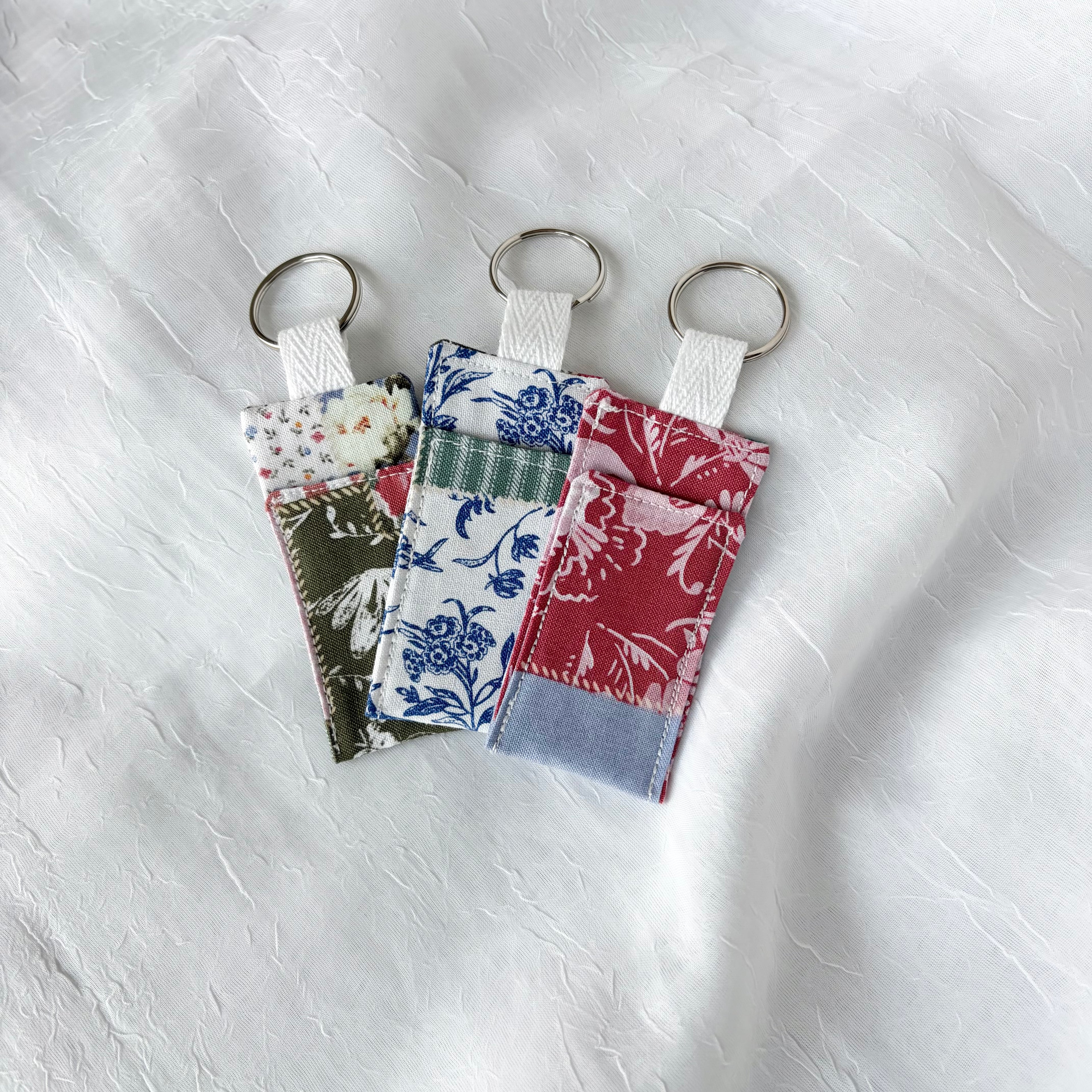 three chapstick holders in colorful patchwork with keychain ring on a textured white background. 