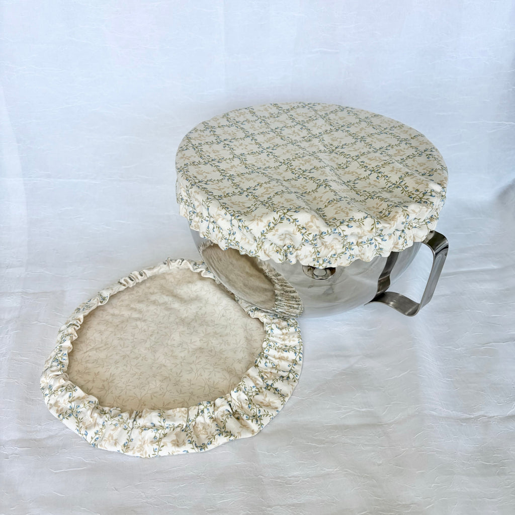 blue, green and cream cover on silver bowl with second cream floral cover next to it on white surface