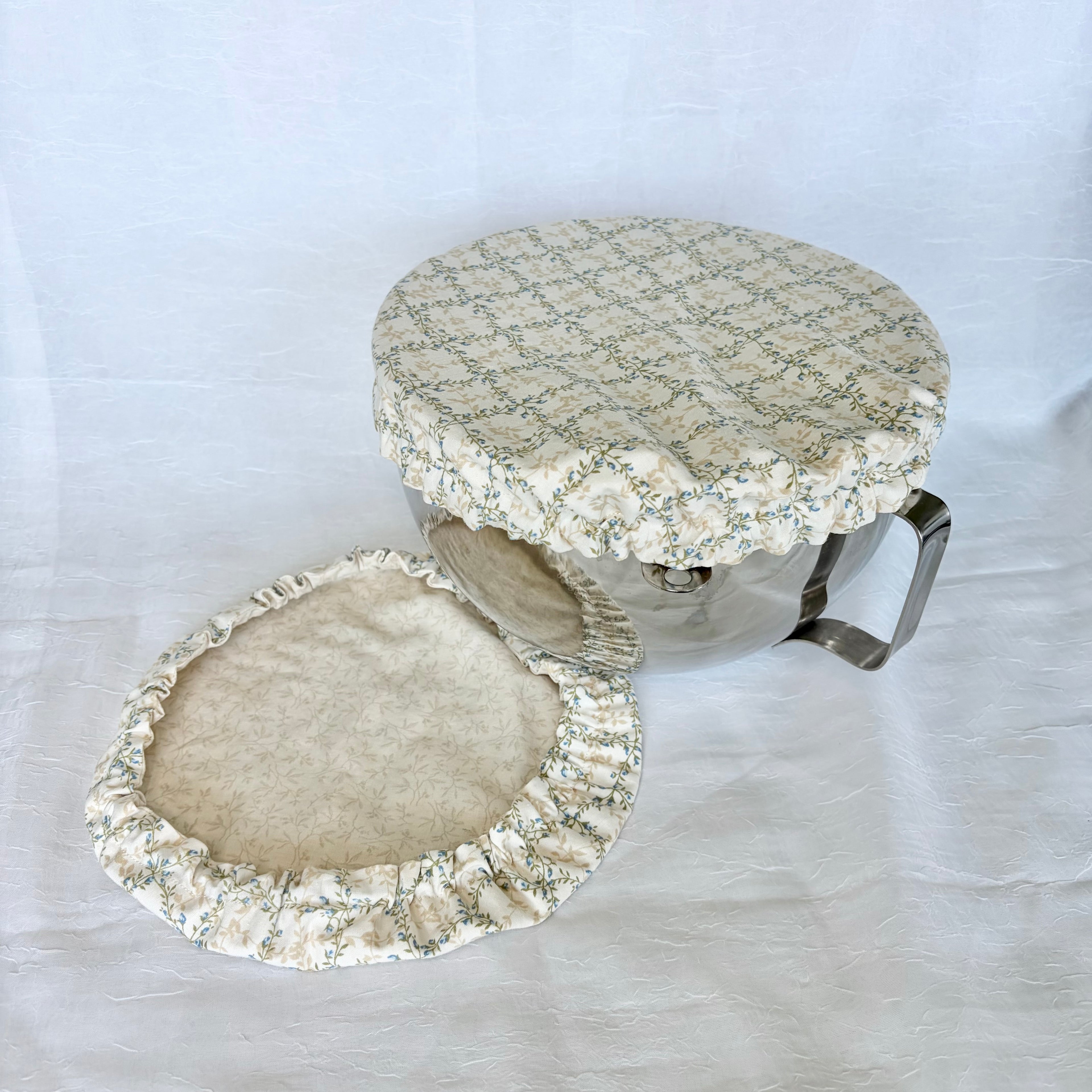 blue, green and cream cover on silver bowl with second cream floral cover next to it on white surface