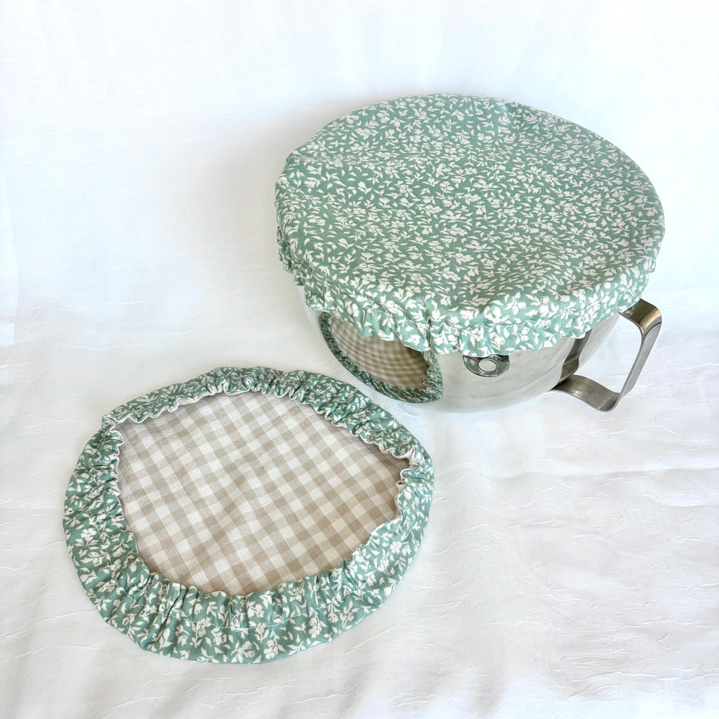 A fabric pot lid cover set with a green and white floral print outer fabric and a beige and white gingham pattern inner lining, shown on a white fabric background.