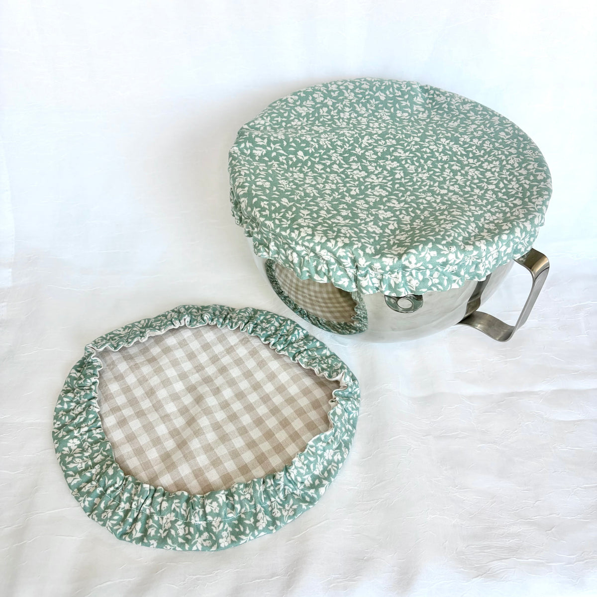 A fabric pot lid cover set with a green and white floral print outer fabric and a beige and white gingham pattern inner lining, shown on a white fabric background.