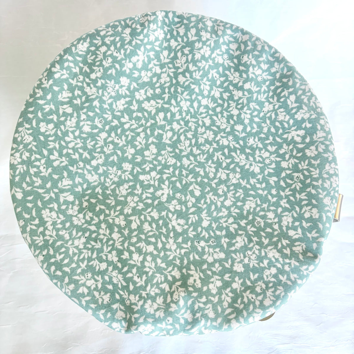 A round fabric-covered bowl with a white floral print on a sage green background, photographed from above on a white fabric surface.