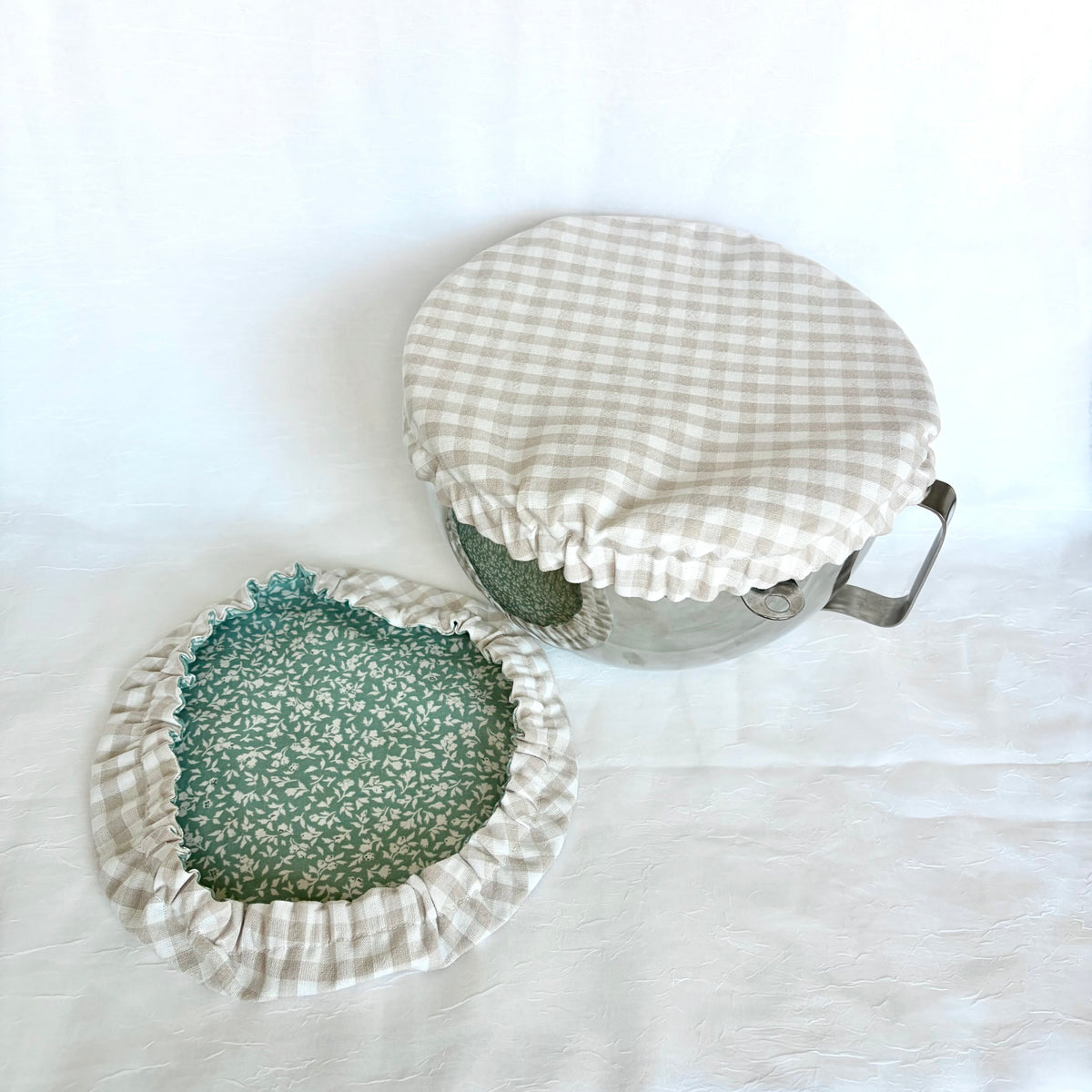 A fabric bowl cover set with a beige and white gingham check pattern and a green floral patterned lining, shown on a white background with one cover on a bowl and another laid flat.