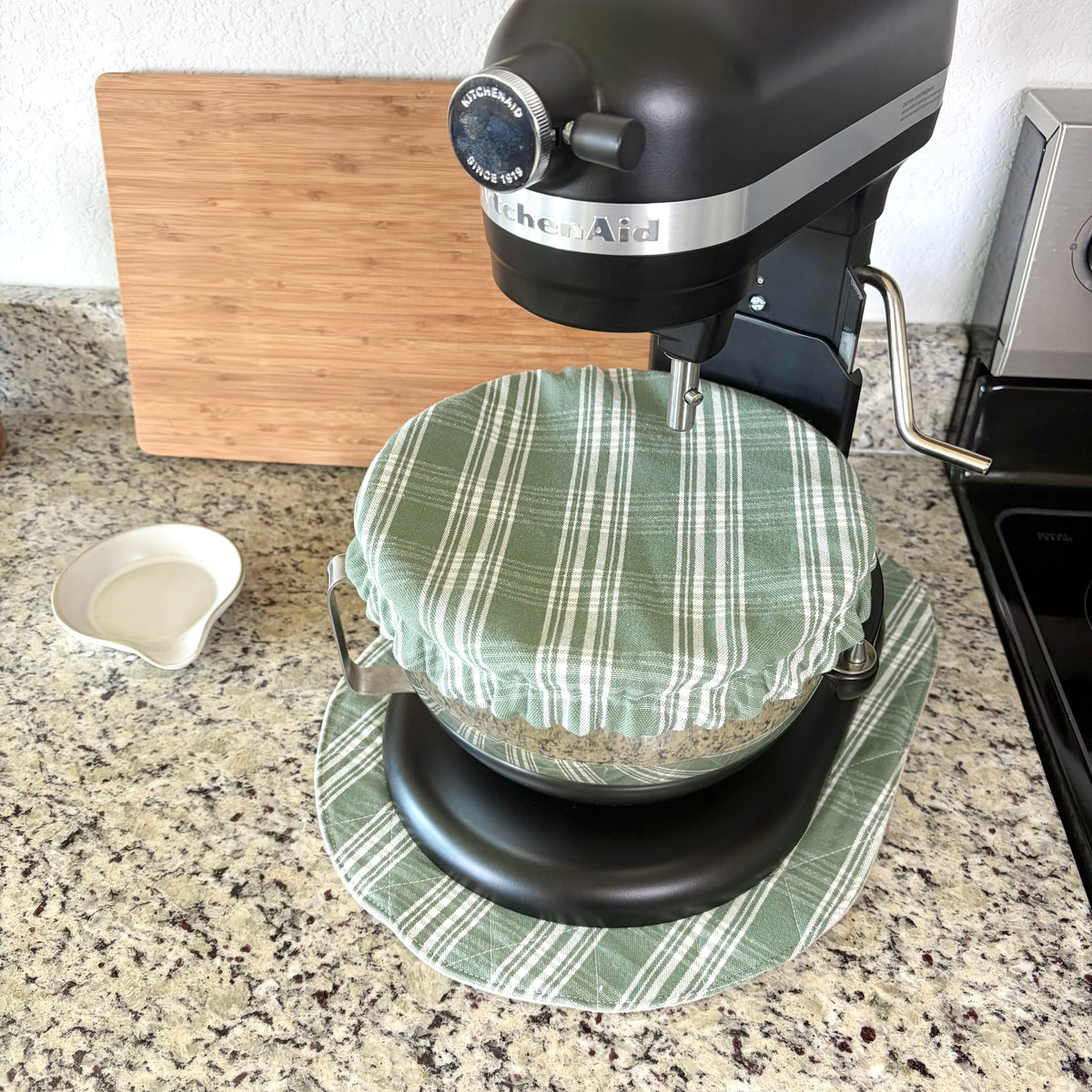 A black KitchenAid stand mixer with a green and white plaid fabric cover on the bowl, set on a matching mat on a granite kitchen countertop with a wooden cutting board and small white dish in the background.