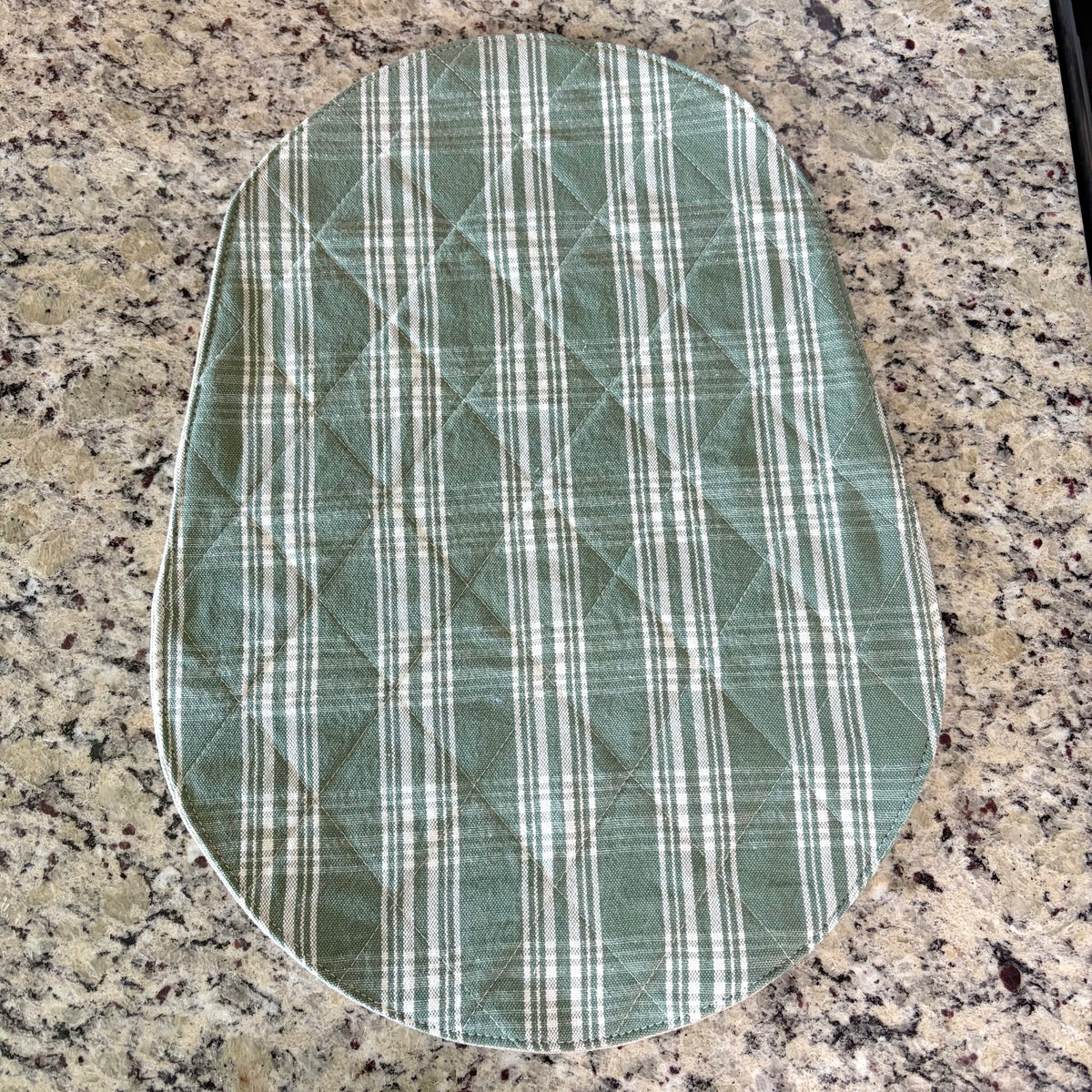 An oval quilted fabric mat, featuring a green and white plaid pattern, laid flat on a speckled granite countertop. Bowl Lift Size