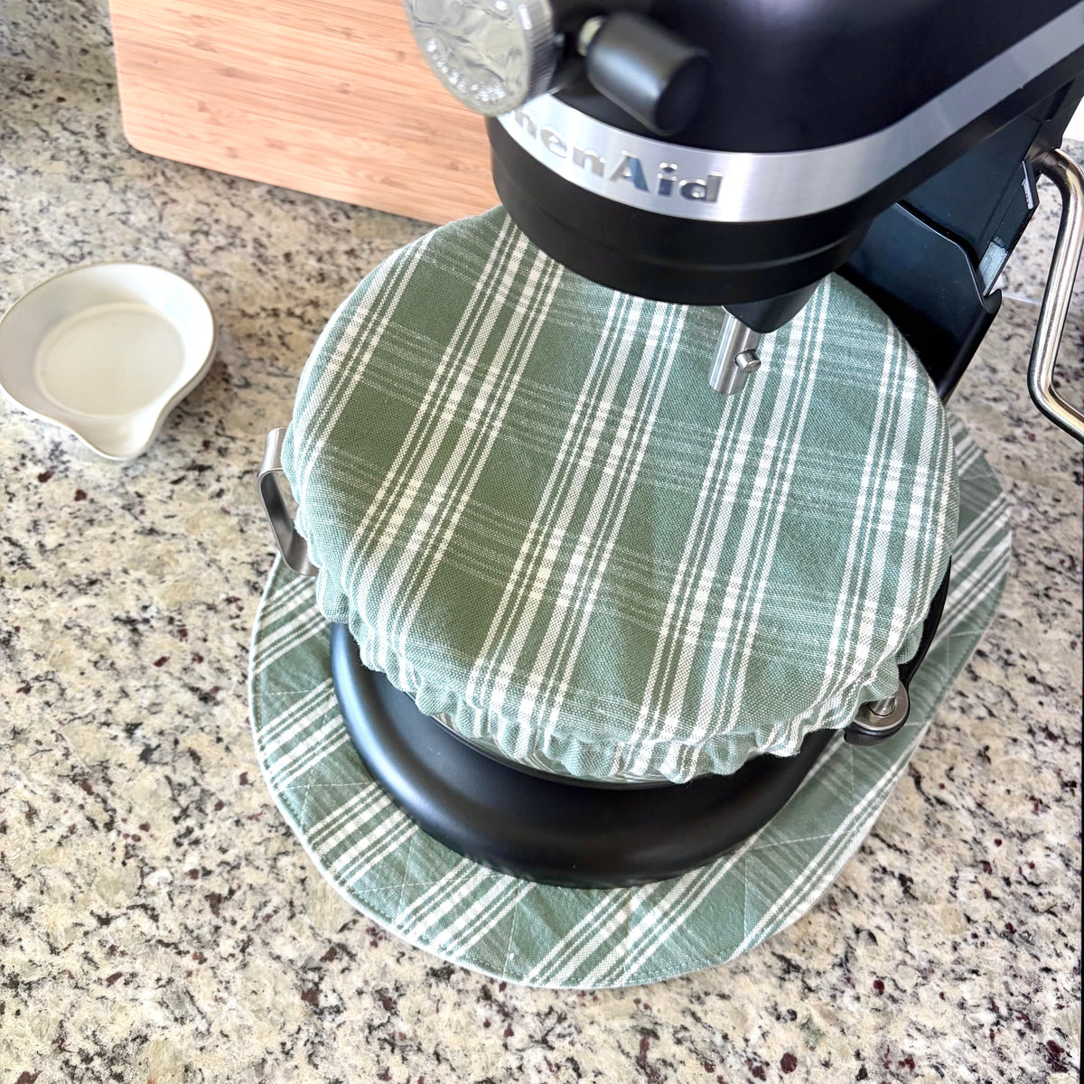 A green and white plaid bowl cover fitted over the lid of a black stand mixer, resting on a granite countertop next to a small white dish.
