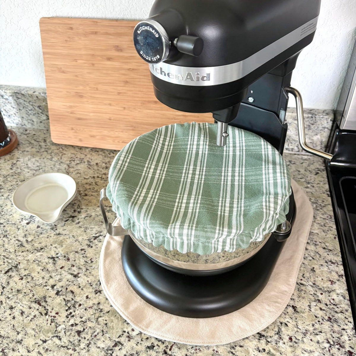 A black KitchenAid stand mixer with a green and white plaid cloth cover on the mixing bowl, sitting on a tan mat on a granite kitchen countertop.