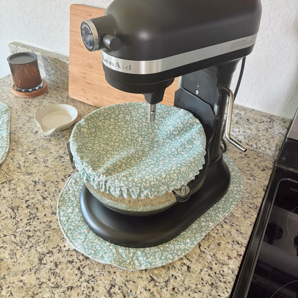 A black KitchenAid stand mixer sits on a granite kitchen countertop, covered with a light green and white floral fabric mat. A matching fabric mat and cover are visible on the counter.