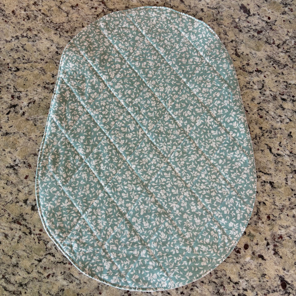An oval-shaped quilted mat with a sage green and white floral pattern, laid flat on a speckled granite countertop. bowl lift size