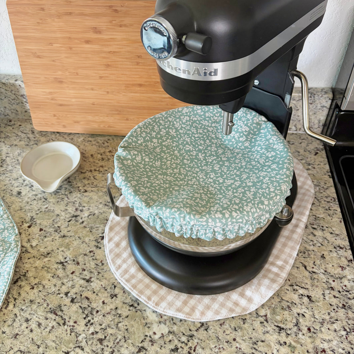 A black KitchenAid stand mixer with a light blue and white floral patterned fabric cover on the bowl, set on a beige gingham mat on a granite kitchen countertop.