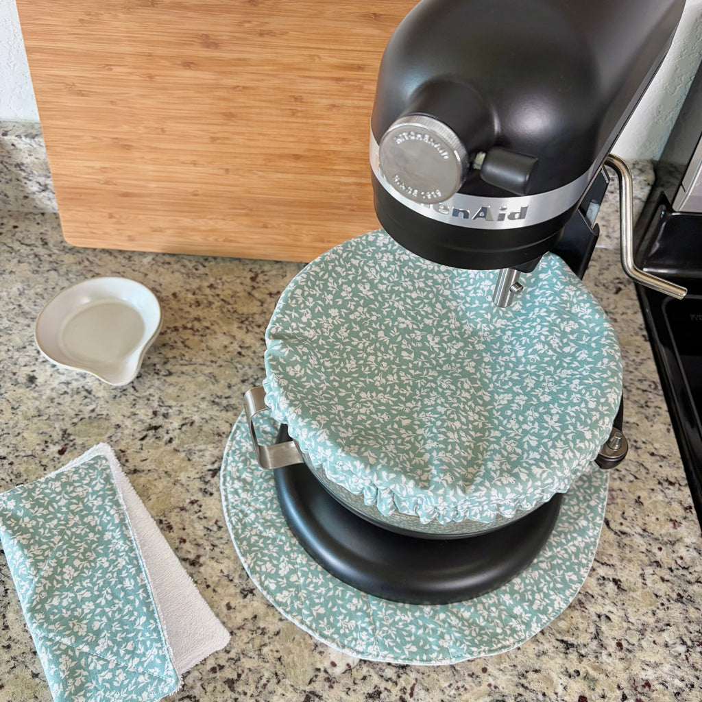 A black KitchenAid stand mixer on a granite countertop with a green and white floral-patterned fabric cover on the bowl. A matching fabric cloth and small white dish are also visible on the counter, with a wooden cutting board in the background.