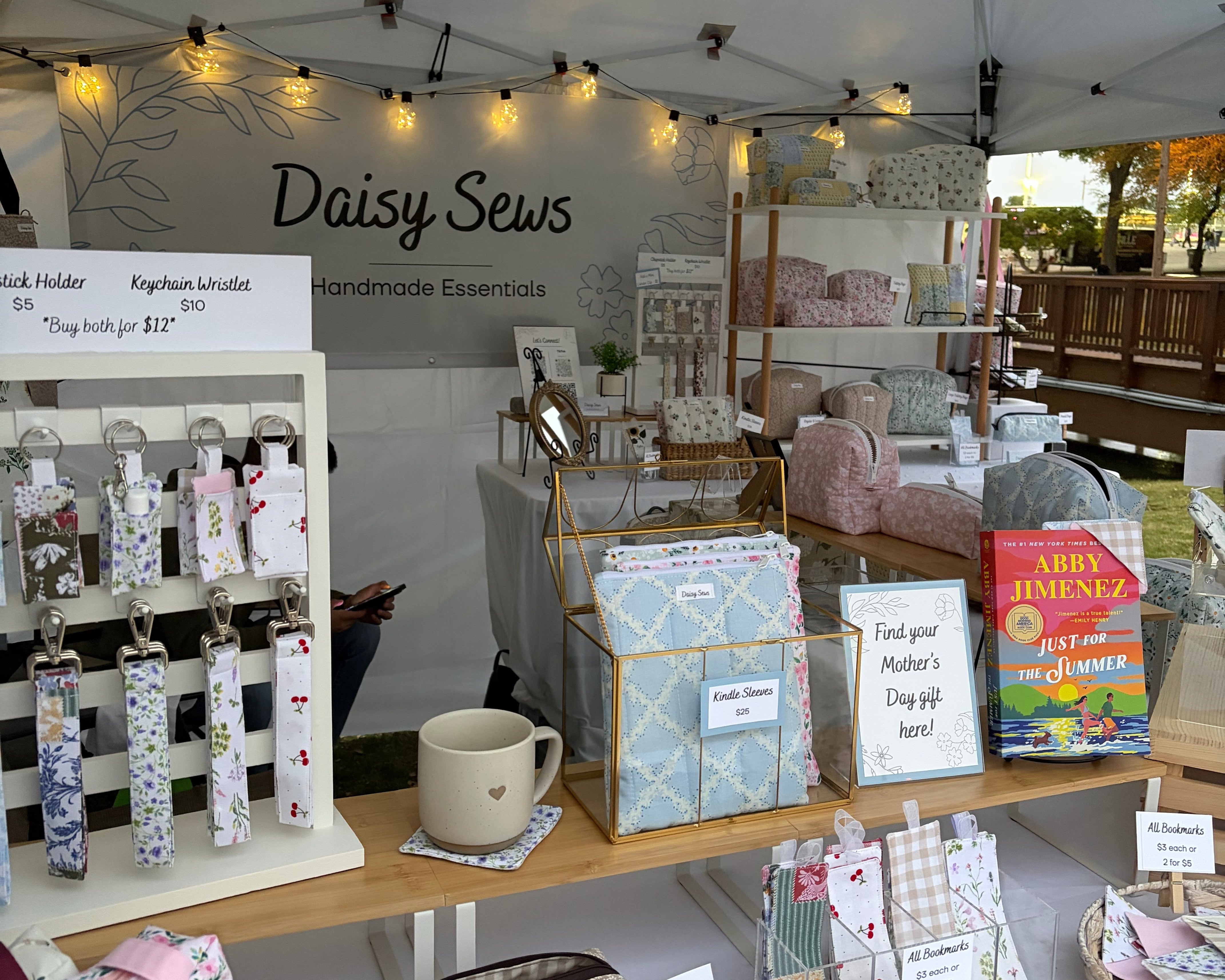 market booth display under a white tent. wooden displays with keychains bookmarks, coaster and toiletry bags in floral prints. A banner saying daisy sews hangs in the background with outdoor lights