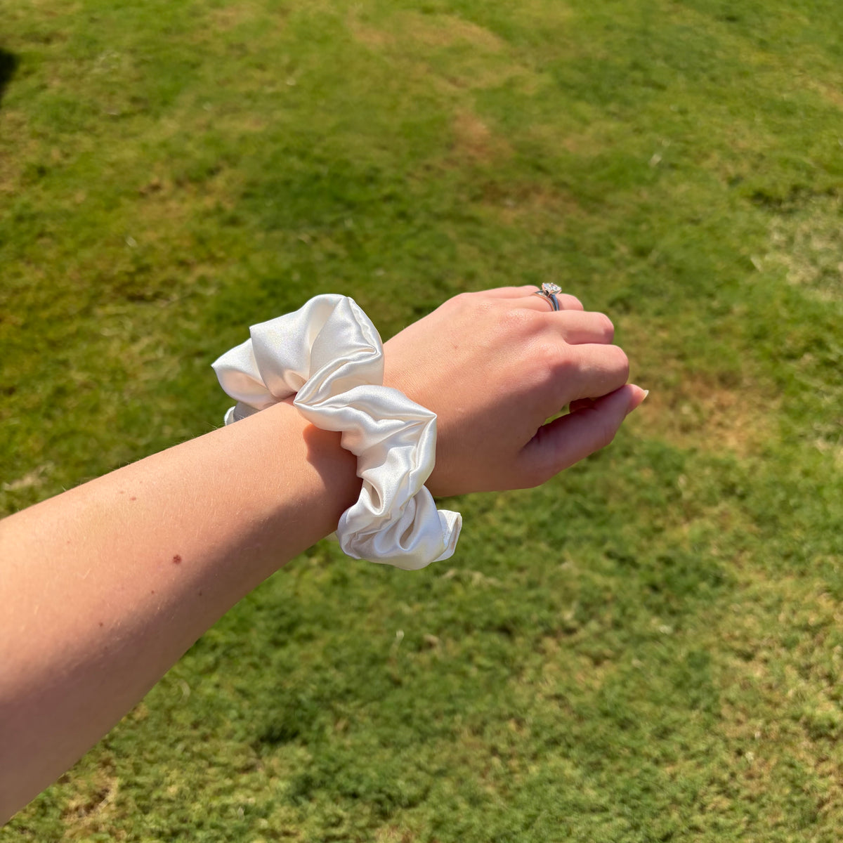 A white satin scrunchie worn on a person's wrist, photographed outdoors against a blurred green grass background.
