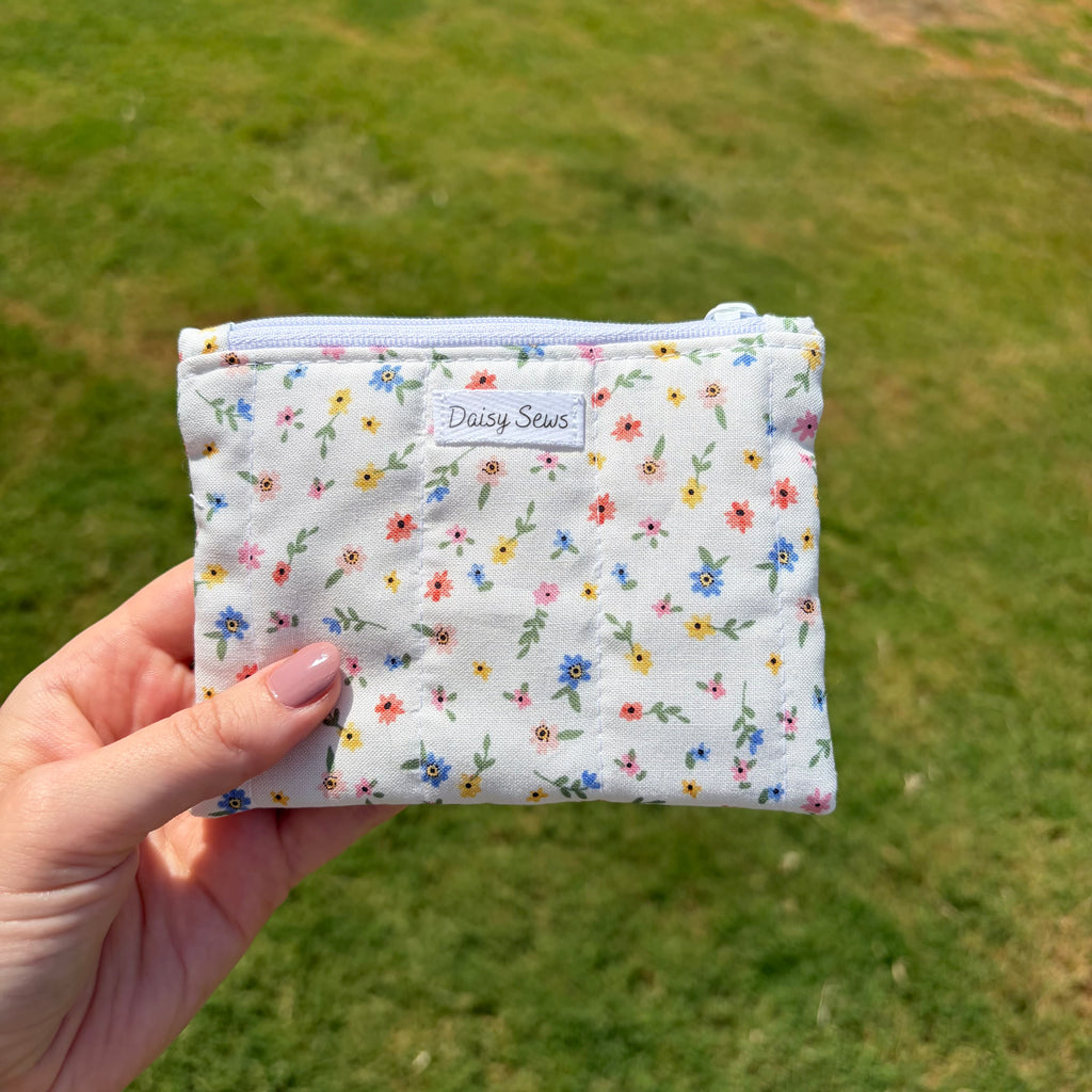 A hand holding a small zippered pouch with a white background and a colorful floral print, set against a blurred green grass background. The pouch has a label that reads 'Daisy Sews'.