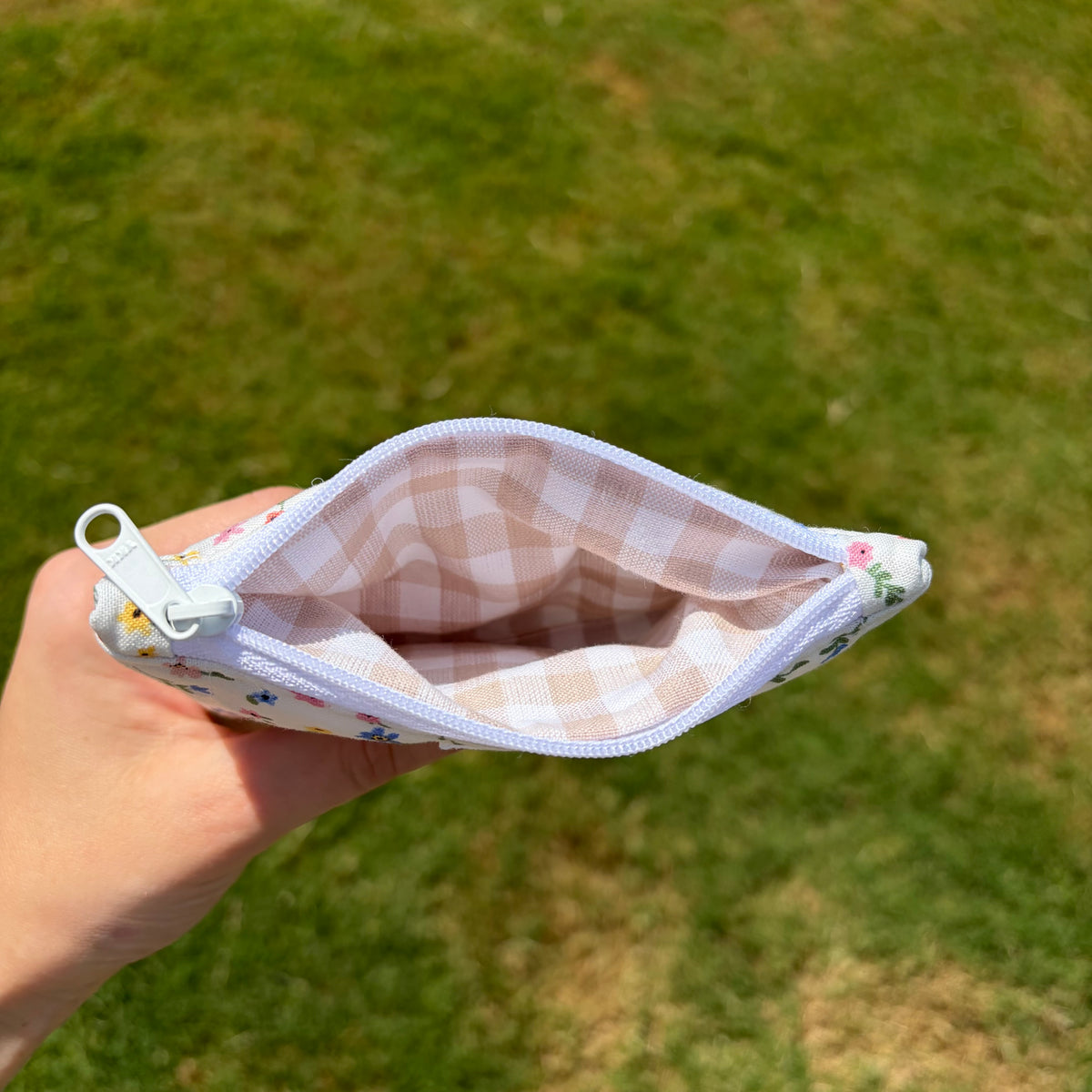 A hand holding an open small zip pouch outdoors over a grassy lawn. The pouch has a white and beige gingham lining, a white zipper, and a white exterior with a small multicolor floral print.