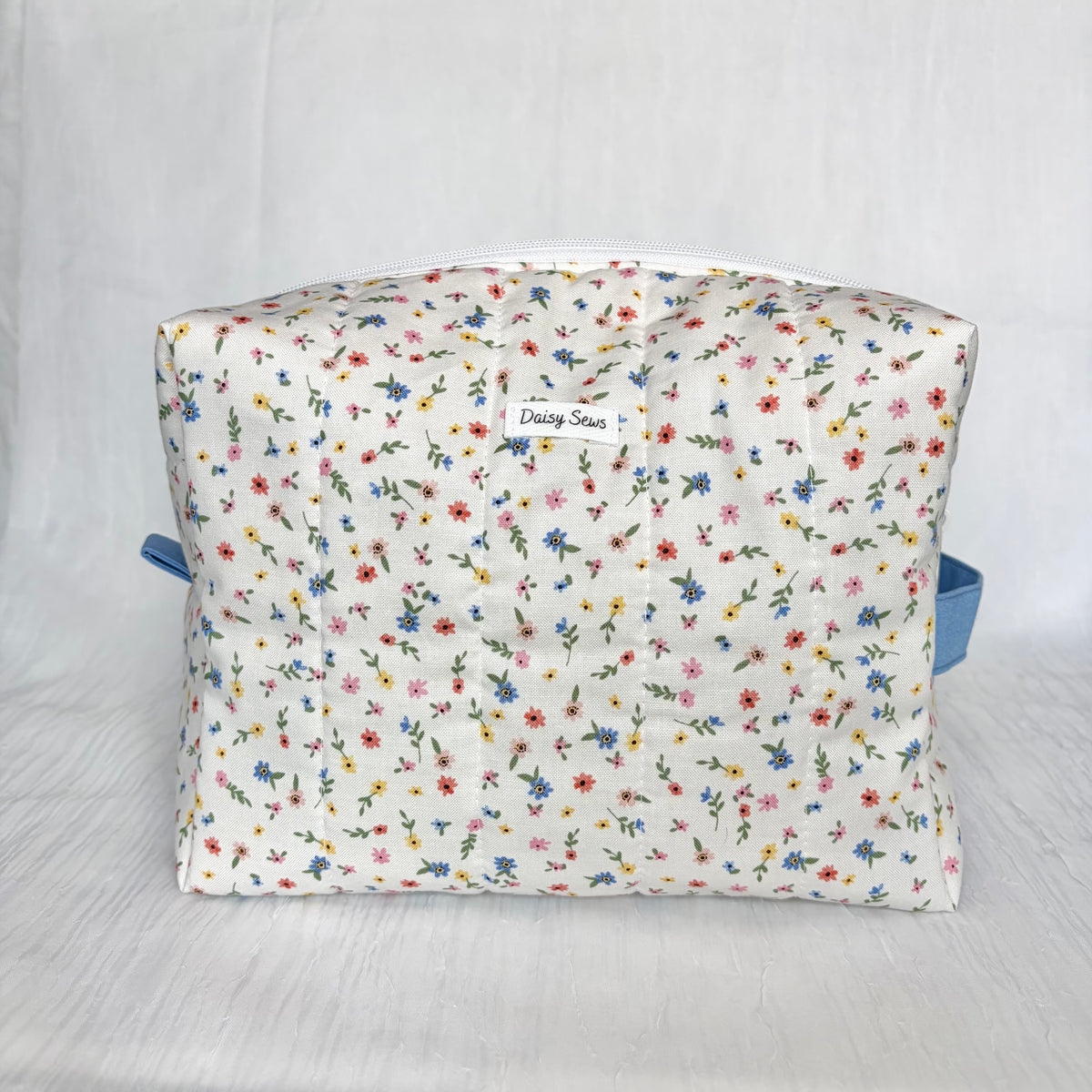 A rectangular toiletry bag with a white background covered in a dense, small-scale floral print of blue, red, pink, and yellow flowers with green stems. It features a small white fabric label in the center that reads 'Daisy Sews' and has light blue side straps, photographed against a white fabric backdrop.
