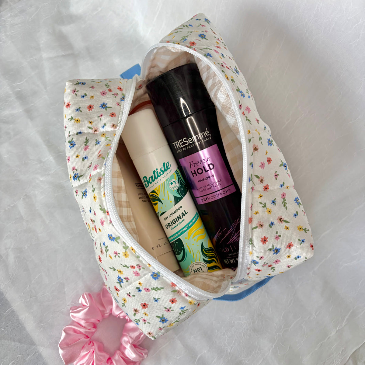 A floral zippered cosmetic pouch filled with toiletries, including a hairspray and a batiste dry shampoo, on a white fabric background.