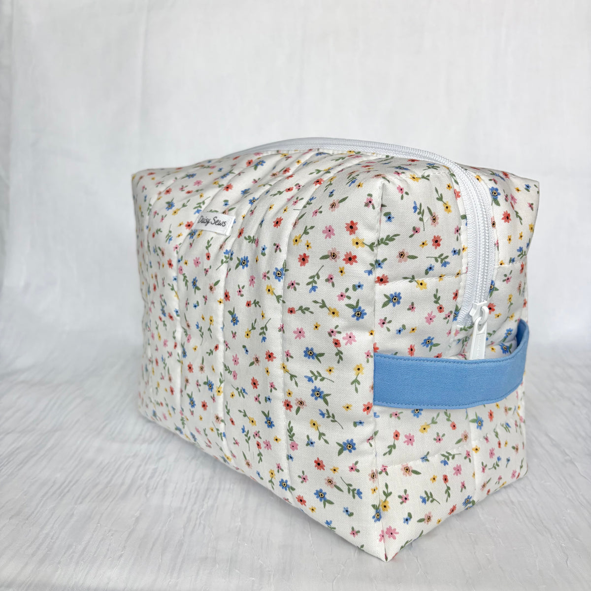 A square quilted fabric toiletry bag with a white background and a colorful floral print, featuring a light blue handle and a white zipper.