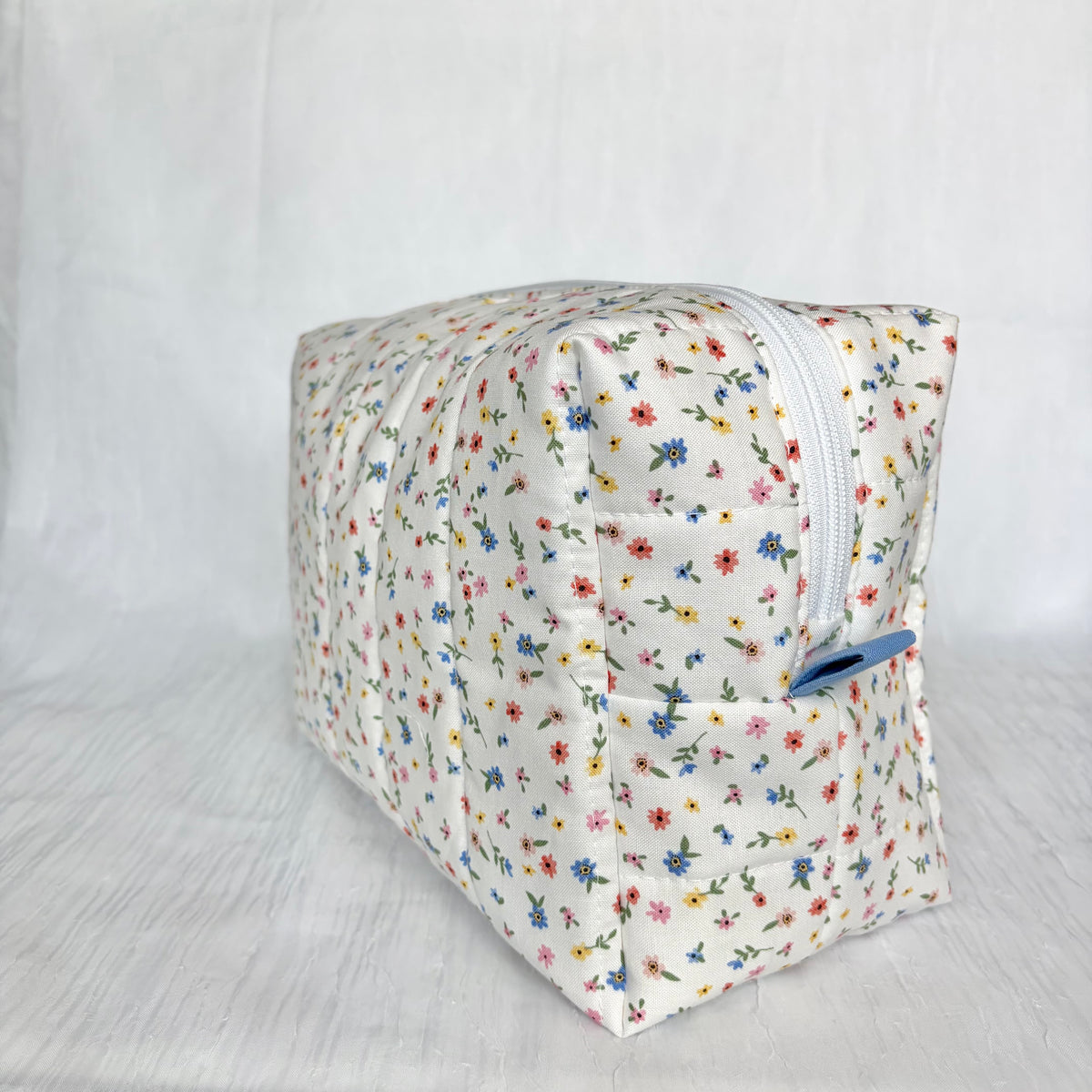 A square zippered fabric pouch with a white background and a small multicolor floral print, featuring a blue pull tab on a white textured fabric backdrop.