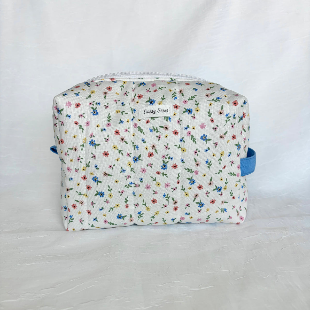 A small rectangular zippered pouch with a white background and a dense multicolor floral print, featuring blue handles on the sides and a small 'Daisy Sews' label on the front.