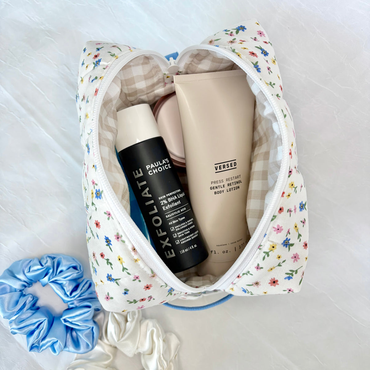 A floral and gingham cosmetic bag is shown open on a white textured surface, containing skincare items including an Exfoliate bottle and a Versed lotion tube, with blue and white scrunchies nearby.