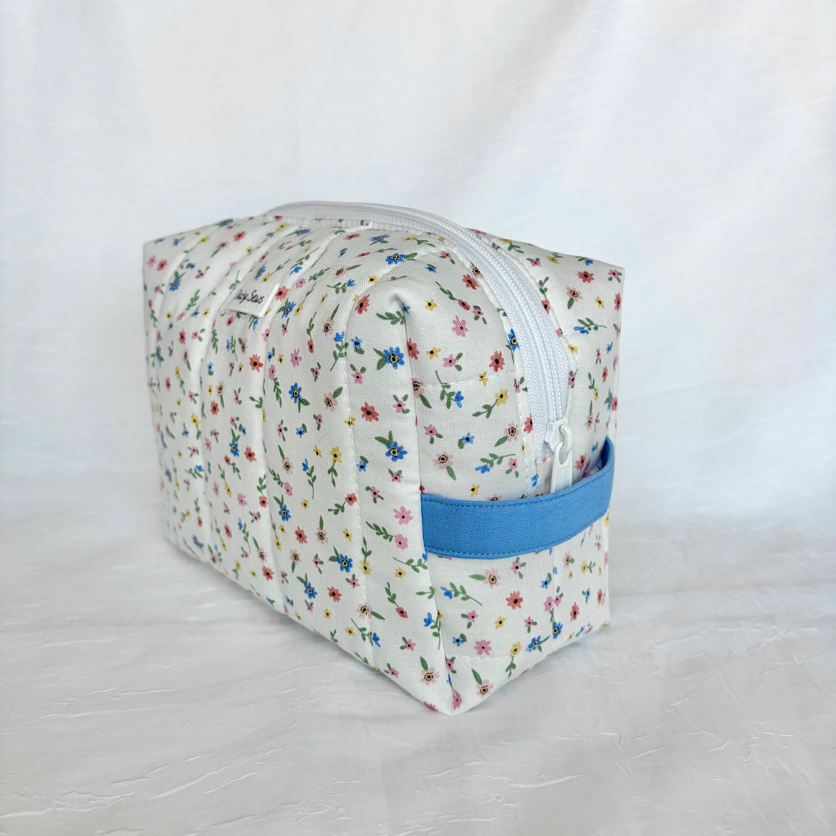 A rectangular fabric zippered pouch with a white background and a colorful floral print, featuring a blue fabric loop handle on the side.
