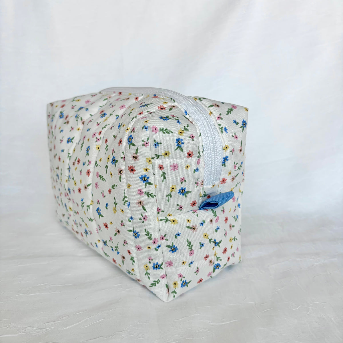 A rectangular zippered pouch with a white background and a colorful all-over floral print, featuring a blue zipper pull, photographed against a white fabric backdrop.