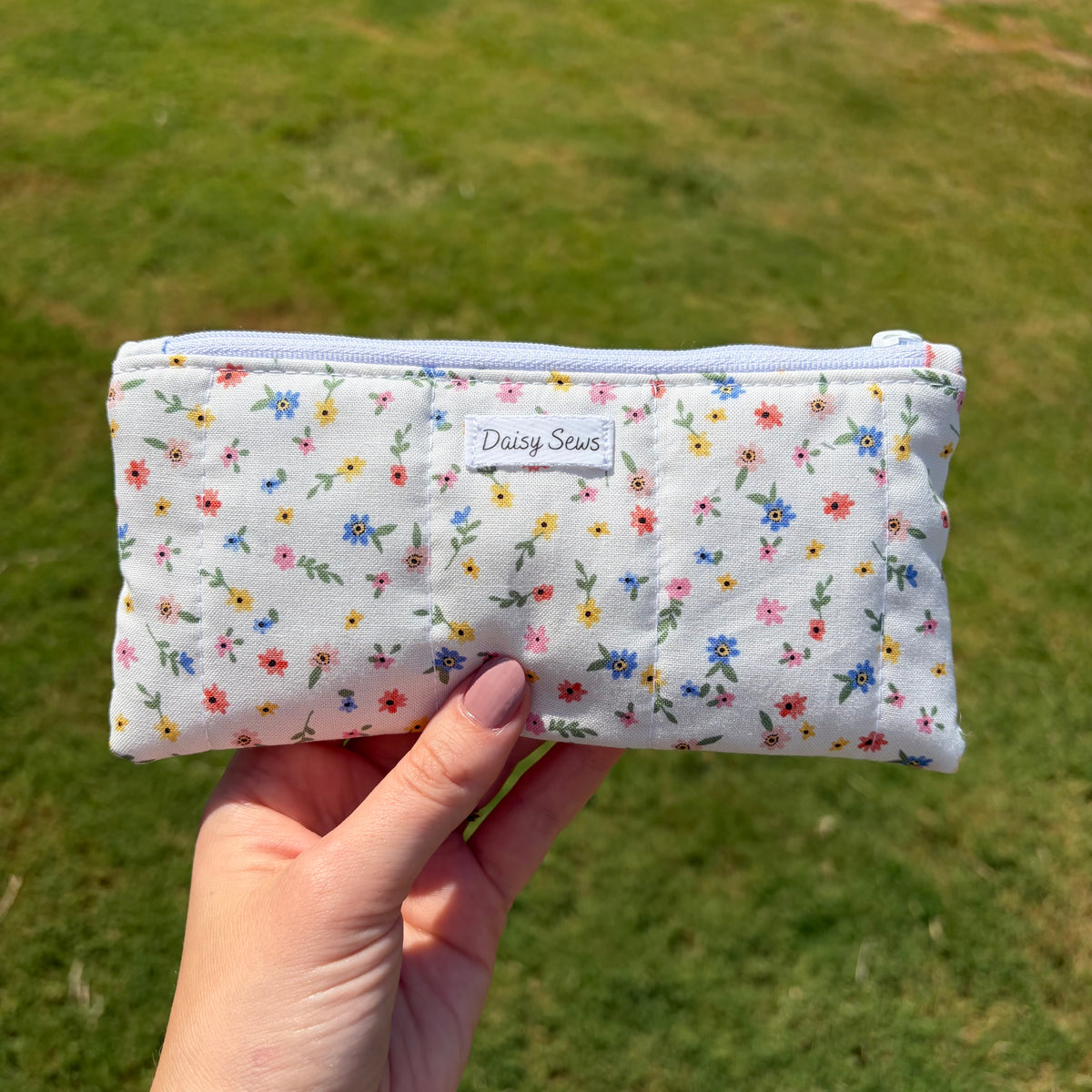 A hand holds a small zippered pouch with a white background and a colorful floral print. The pouch has a small fabric label that reads 'Daisy Sews' and is photographed outdoors against a blurred green grass background.