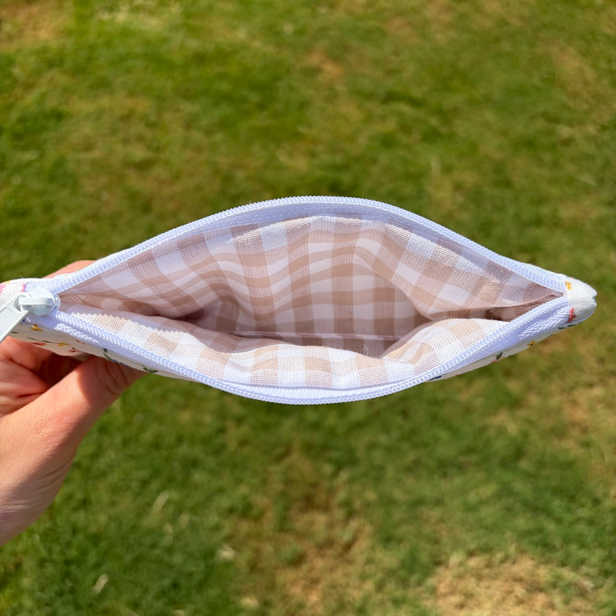 A hand holding open a small zip pouch outdoors, showing a beige and white gingham pattern lining against a blurred green grass background.
