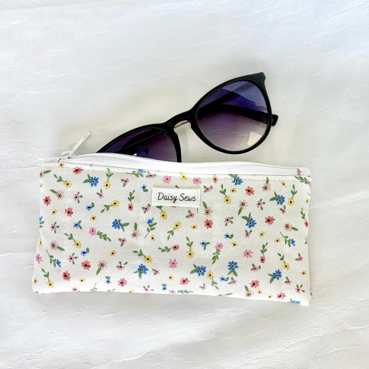 A white zippered pouch with a colorful daisy floral print, featuring a small white label that reads 'Daisy Sew'. Black cat-eye sunglasses are resting on top of the pouch, which is laid flat on a textured white fabric background.