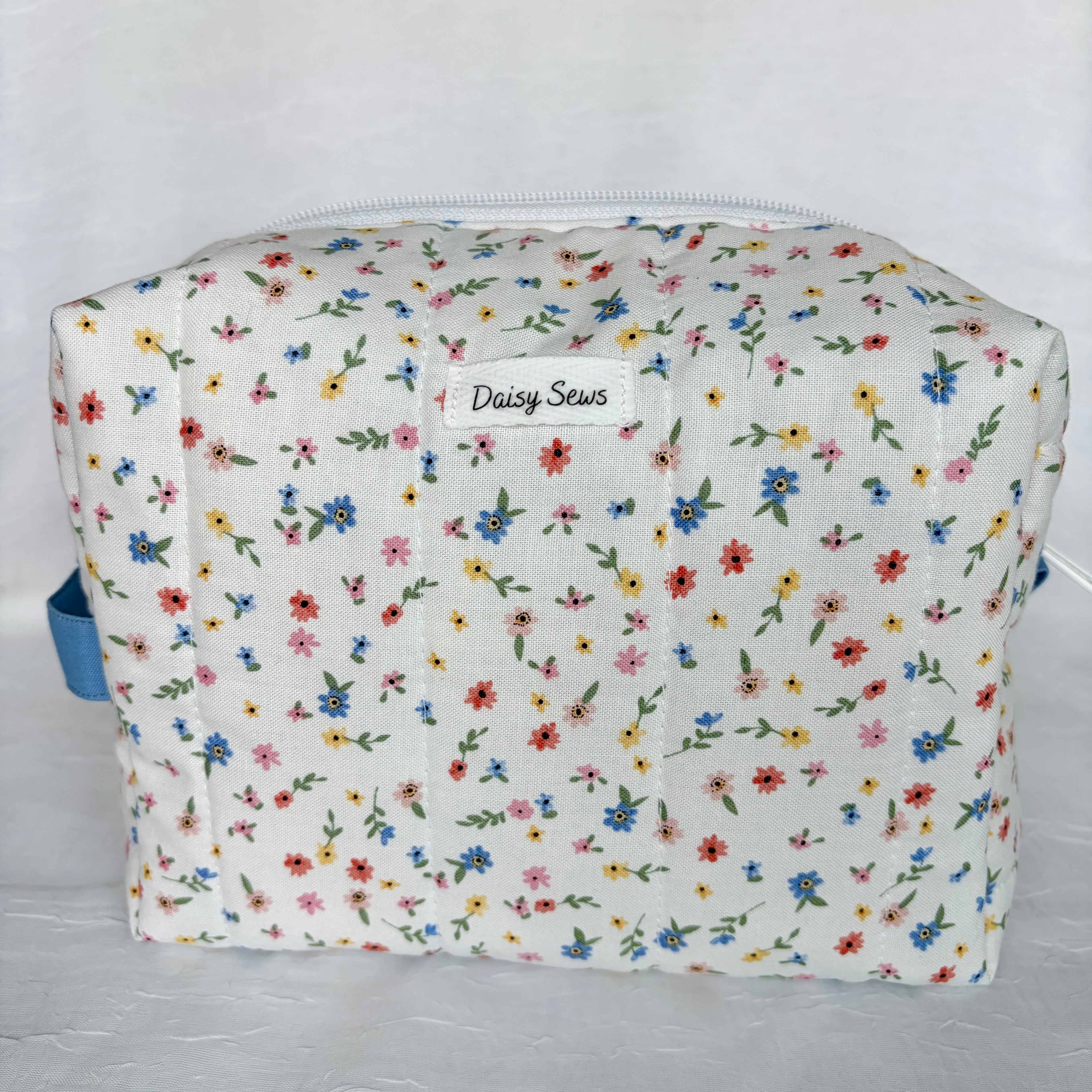 little blue, yellow, pink and red flowers with green leaf patterned boxy bag with white zipper/label and blue handle on white background