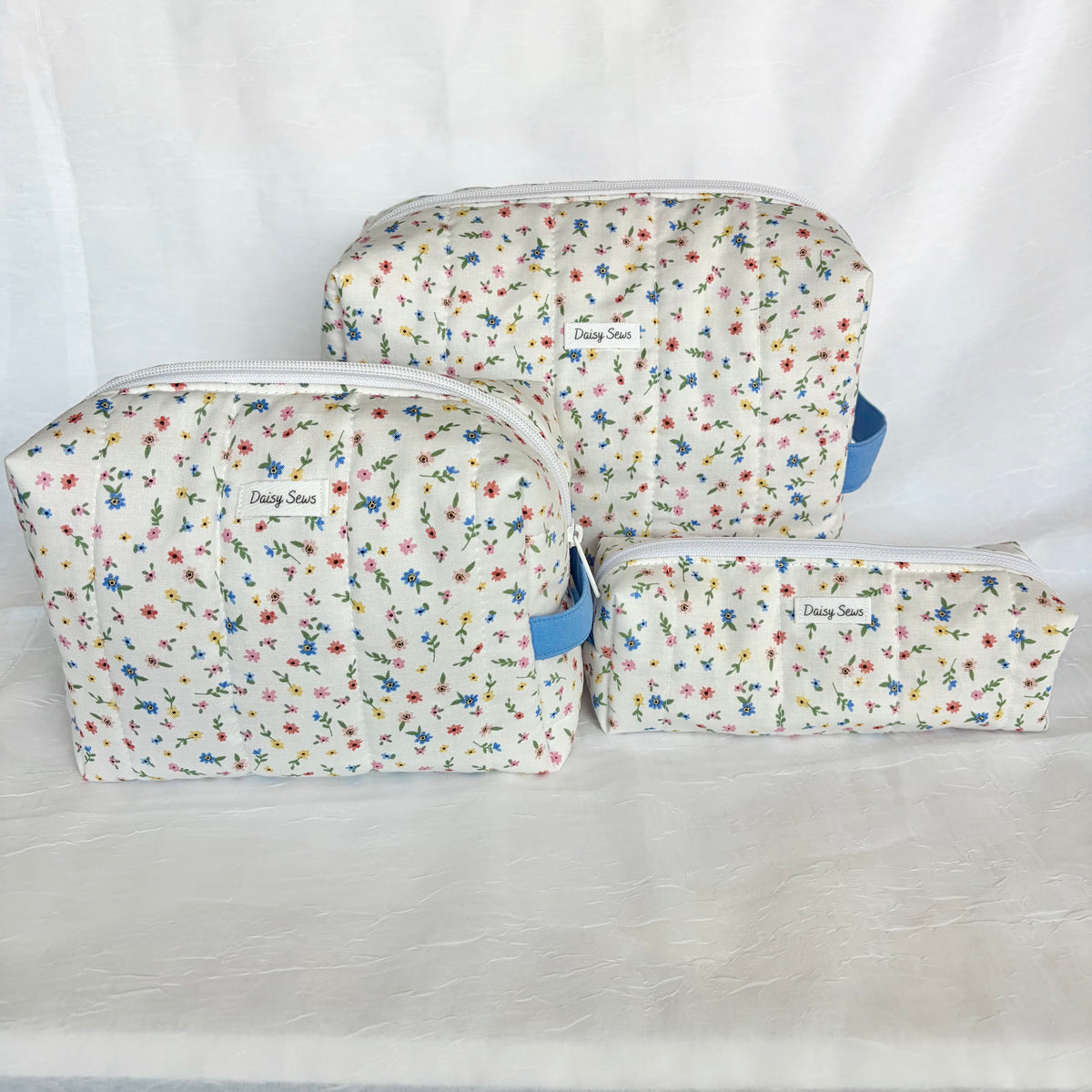 Three fabric zippered pouches in different sizes with a white background and a delicate multicolor floral print, featuring blue side straps and white labels.