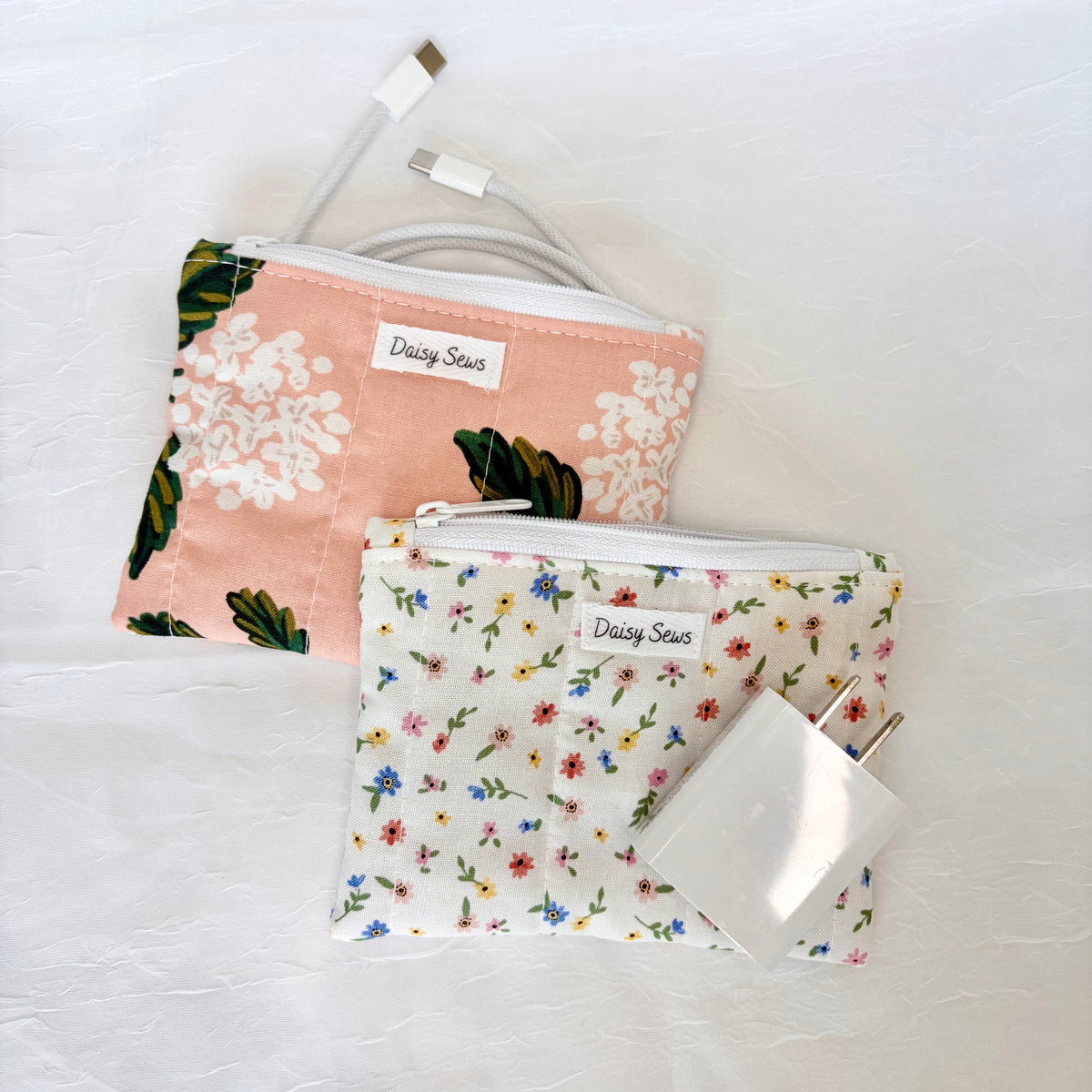 Two fabric zippered pouches by Daisy Sews displayed on a white textured background, with a white USB-C cable and a white power adapter resting on top of them. One pouch is light pink with white floral print and green leaves, and the other is white with a dense multicolor floral print.